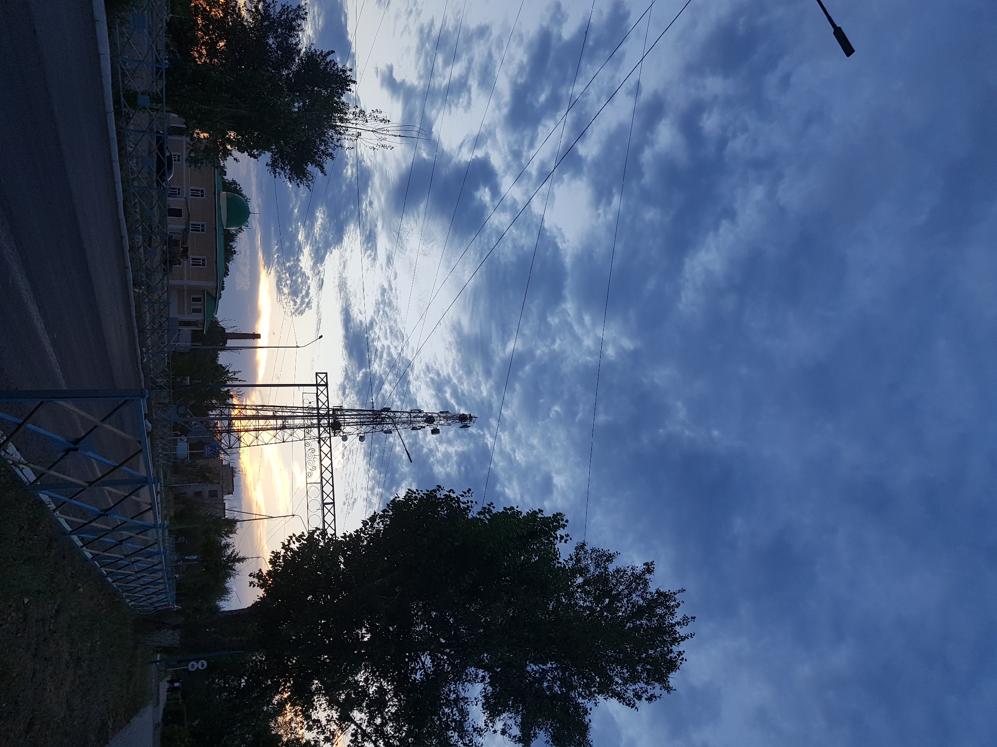The image depicts a scene in the city of Kurchatov, Kazakhstan, during what appears to be dusk. A street runs horizontally across the lower portion of the frame, with a blue and white patterned metal fence running along its right side. Beyond the fence, there is a sidewalk with grass and trees.
Dominating the left side of the background is a building with a green domed roof, likely a mosque, with other buildings and trees visible behind it. To the right of the mosque, a tall transmission tower with multiple antennas is visible, silhouetted against the sky. A large tree with dark foliage stands prominently in the mid-right of the image, partially obscuring the transmission tower and the sky.
The sky is filled with dark blue and gray clouds, with a hint of orange and yellow sunset peeking through the clouds in the distance, behind the transmission tower. Power lines crisscross the sky, and a lamppost is visible on the far right. A dark-colored car is parked in front of the building on the left. The overall atmosphere is twilight, with artificial lights not yet fully illuminated.
