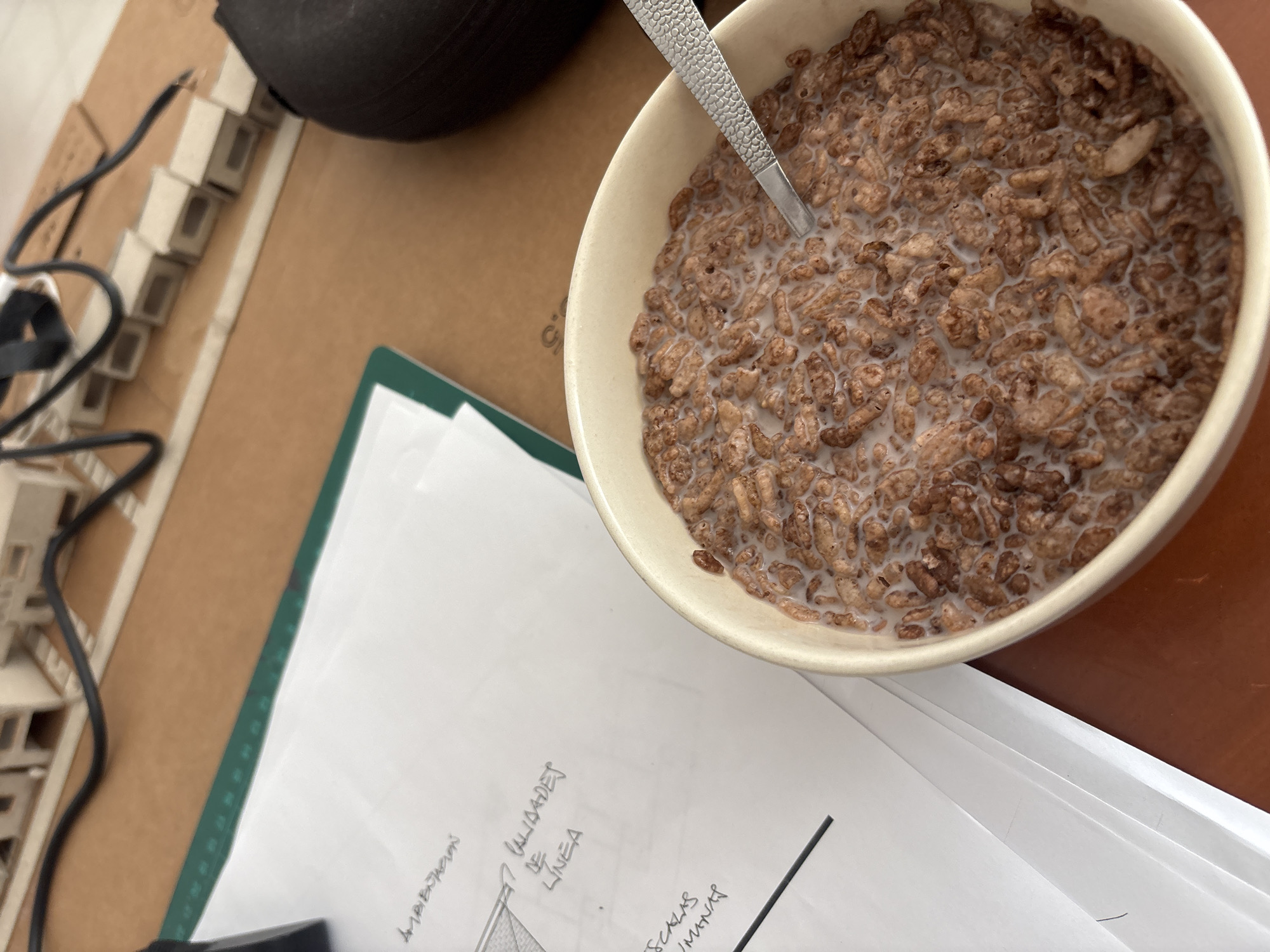 A high-angle shot shows a bowl of cereal with milk on a brown surface next to architectural drawings and a small model. The cereal is chocolate-flavored and a silver spoon is visible in the bowl. The drawings are on white paper placed on a green cutting mat, with some text in Spanish, "VALIDADEZ DE LINEA", and "AMBITOS…". The model is small, made of white material, and has a series of small rectangular shapes connected by wires. A black, round object is above the bowl. The scene is likely indoors, but the specific location in San Pablo del Monte, Mexico cannot be determined without further information.