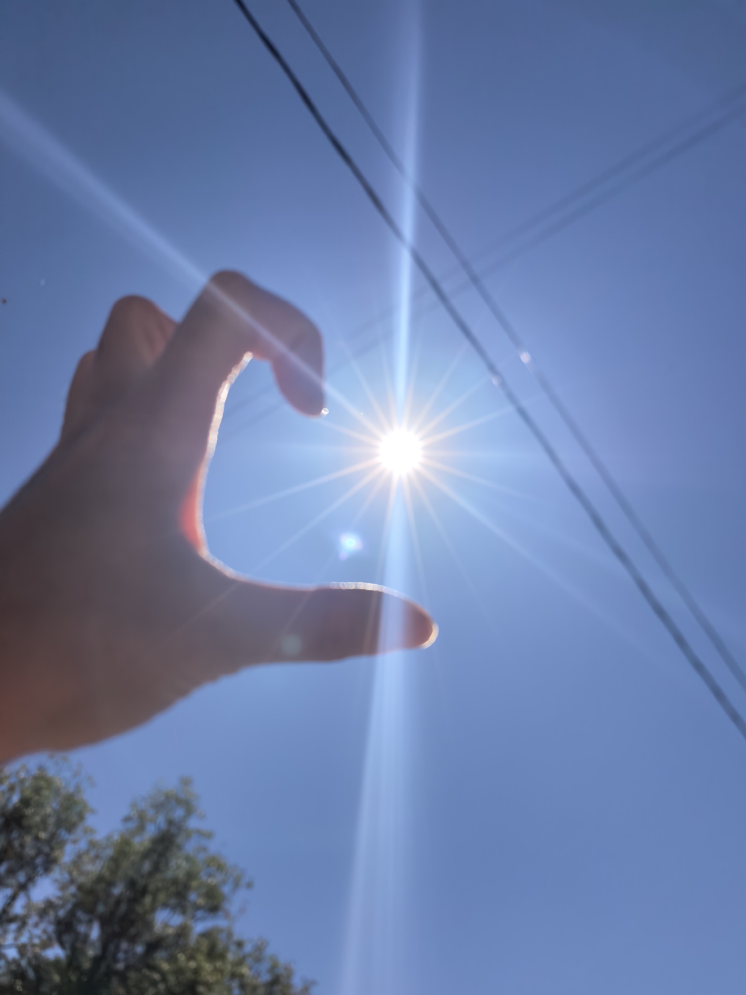 A hand is held up towards the sun against a bright blue sky, creating the illusion of holding the sun between the thumb and forefinger. Power lines run diagonally across the top right portion of the frame. The bottom of the frame shows a glimpse of trees with green foliage. The location is not discernible within the image; therefore, a description related to the city of Sterlitamak, Russia, cannot be provided.