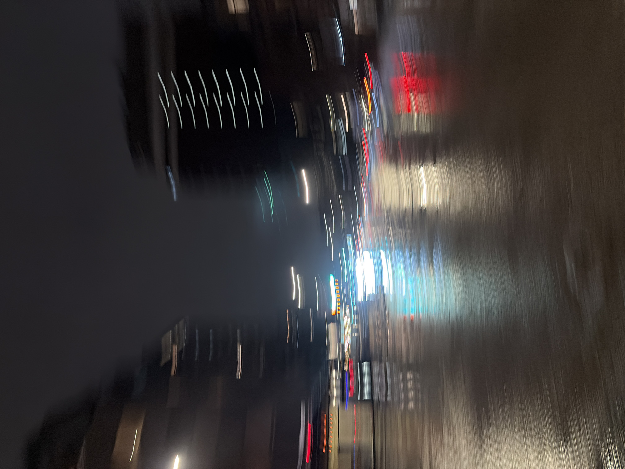 The image is an abstract, motion-blurred shot of a street scene at night. Tall buildings are visible on either side of the street, with lights streaking vertically due to the long exposure or camera movement. The lights suggest the presence of cars and streetlights. The location is possibly in Montreal, Canada, suggested by the urban setting and building styles. The ground appears to be wet, reflecting the lights. No people are clearly visible due to the motion blur. The overall impression is one of a bustling urban environment at night, captured in a dynamic and abstract way.