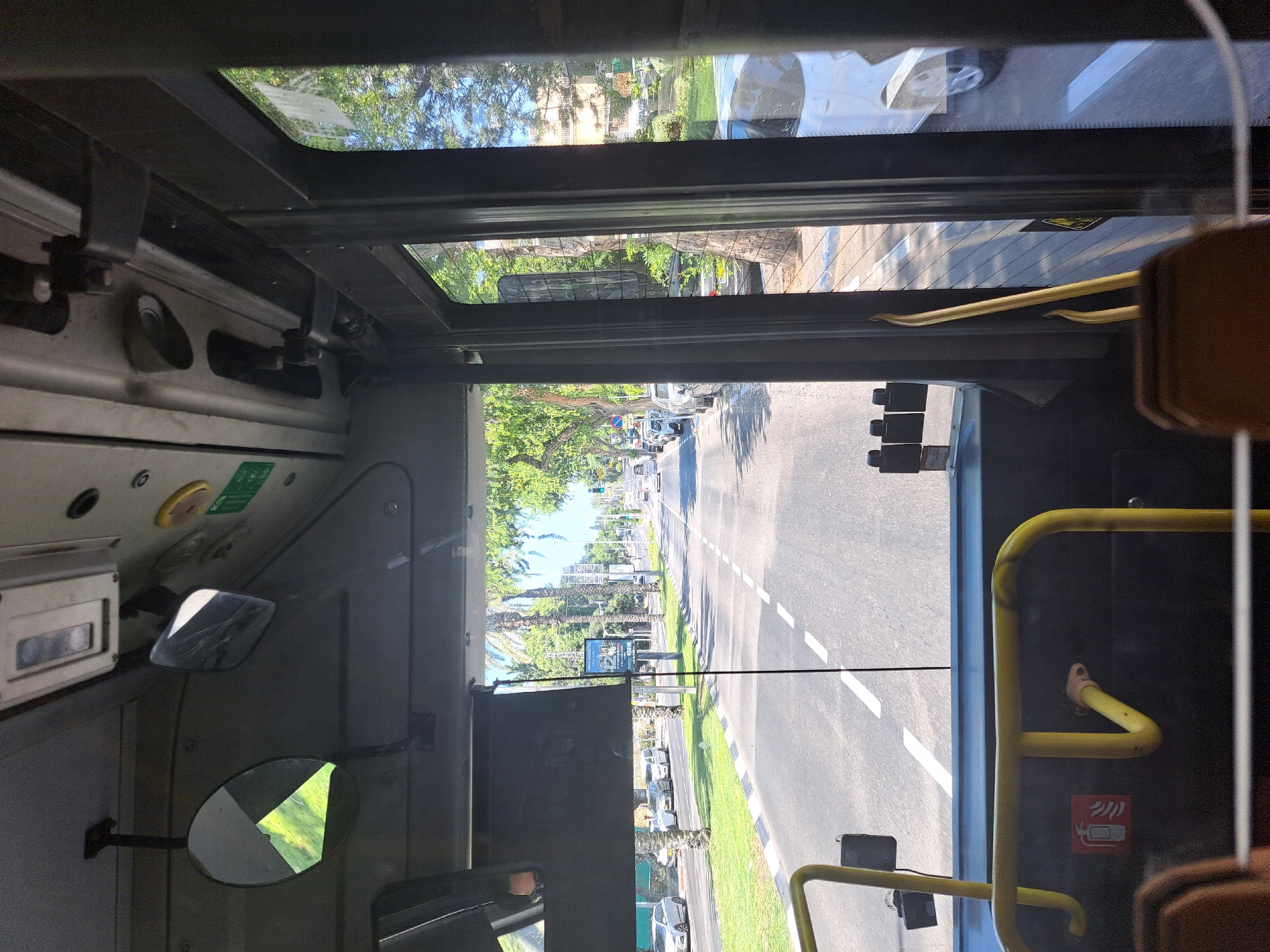 The image is taken from the interior of a bus in Raanana, Israel, looking forward through the windshield. A road with two lanes in each direction is visible, lined with trees and palm trees. Several cars are driving on the road, and parked cars are visible on the side. There are streetlights, traffic lights, and signs along the road. Buildings and residential areas can be seen in the background. The interior of the bus is visible in the foreground, with a yellow handrail, a fire extinguisher sign, and a seat.