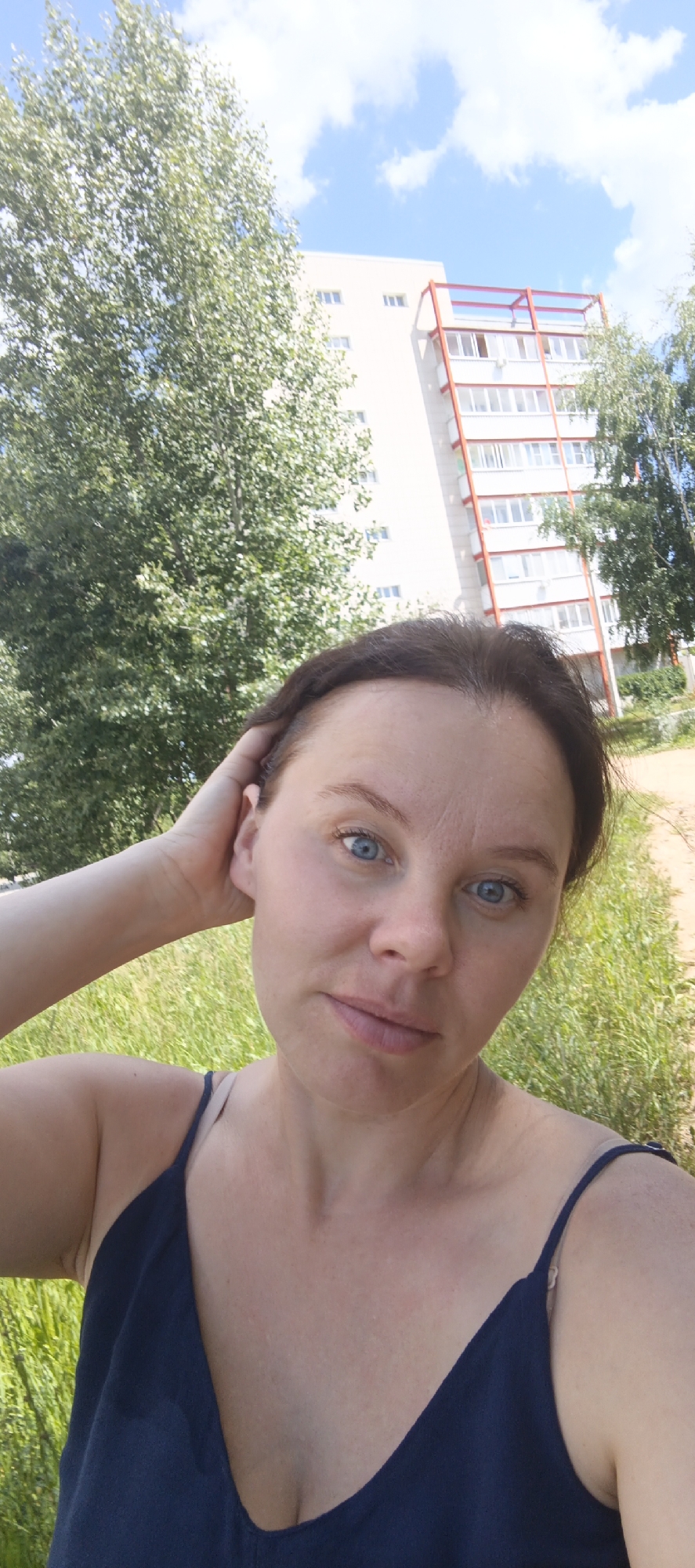 This is a selfie of a woman with blue eyes and brown hair, taken outdoors. She is wearing a dark blue tank top. Her left hand is resting on her head. In the background, there is a tall apartment building with red accents and balconies, and trees with green foliage. The sky is blue with white clouds. The setting appears to be a residential area on a sunny day, possibly in the afternoon. The woman has a relaxed expression. There is no visible text in the image. The context provided suggests the location is Chaikovsky, Russia.