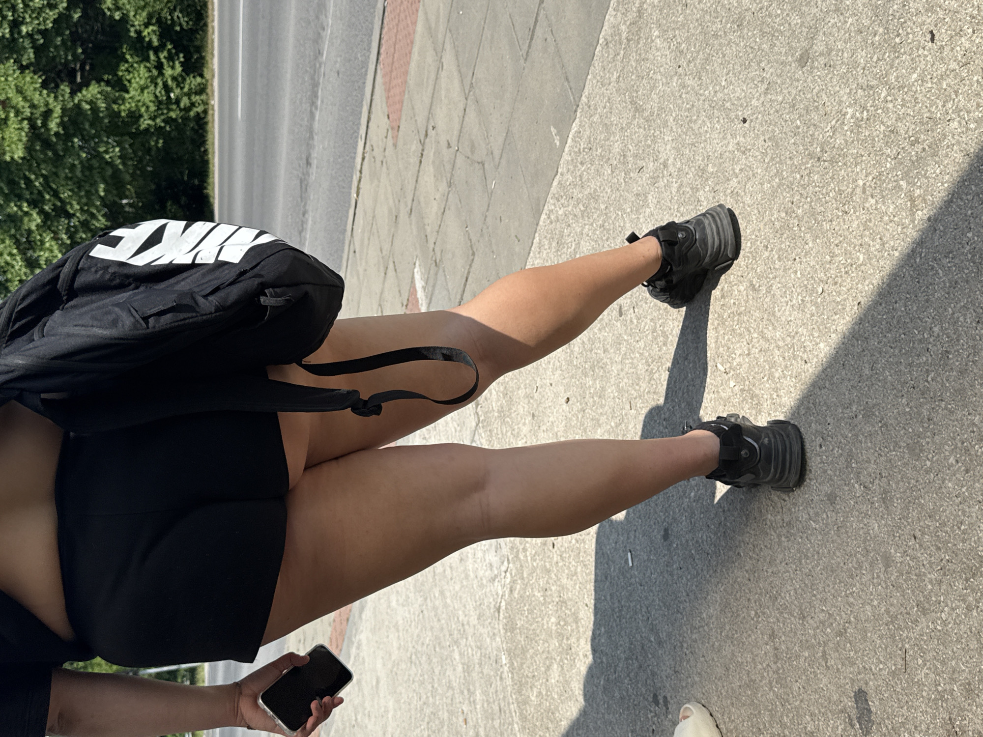 An individual is depicted from the lower back to the feet, walking on a concrete sidewalk during daylight hours in Unknown, Unknown. The person is wearing black athletic shorts and black athletic sneakers. A black backpack, featuring a large white "NIKE" logo, is carried on the left side of the body. A black smartphone is held in the individual's right hand. The skin tone visible on the legs and arm is light brown.
The sidewalk consists of grey concrete pavers, with some reddish-brown sections. A white cigarette butt is visible on the sidewalk surface. On the far left, a portion of a white slipper is present. Adjacent to the sidewalk, an asphalt road with white lane markings extends into the background. A line of green deciduous trees borders the far side of the road. Strong shadows are cast on the sidewalk, indicating clear, sunny weather.