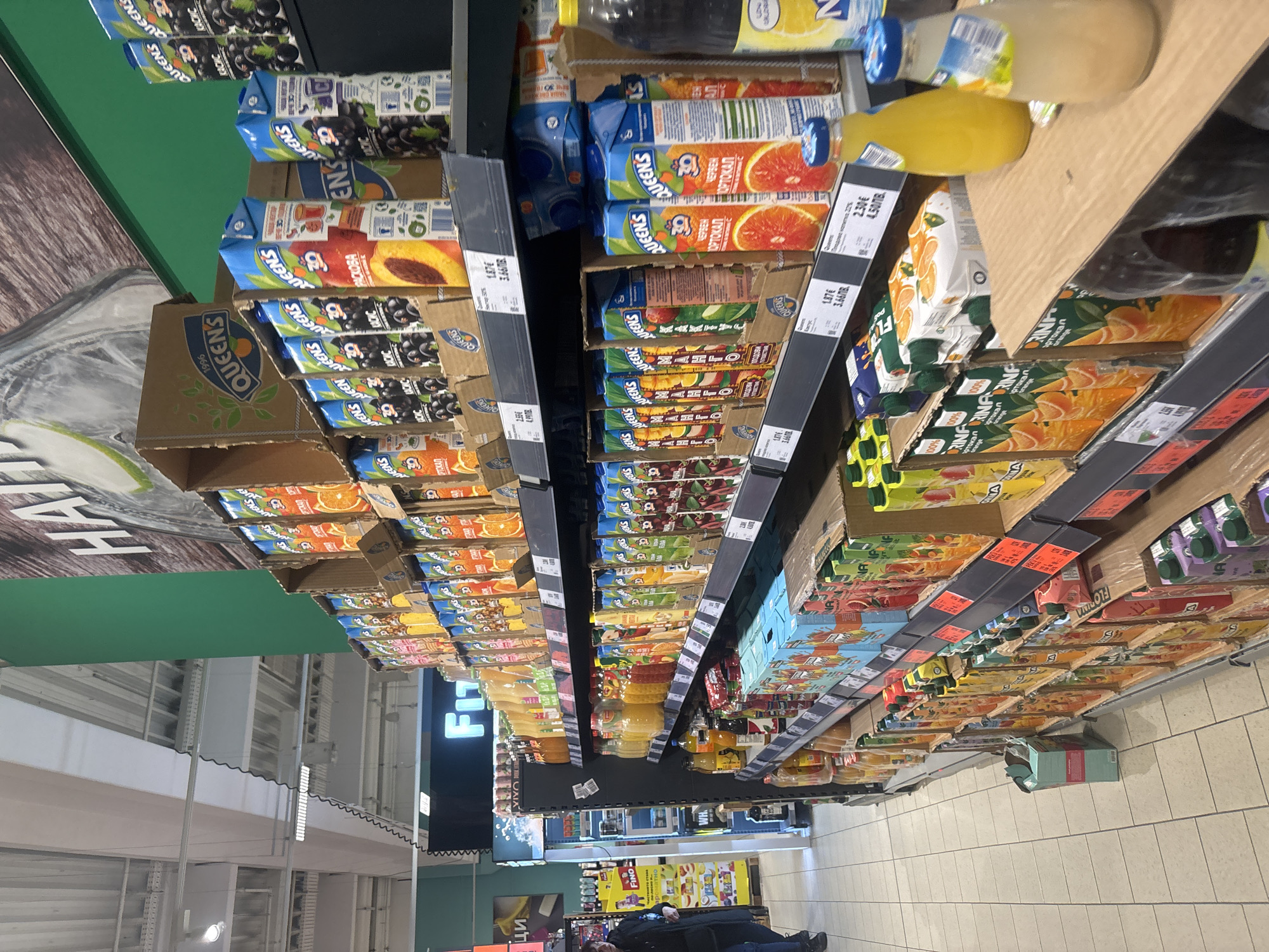 The image shows a supermarket aisle in Plovdiv, Bulgaria. Shelves are stocked with juice boxes and bottles, primarily the "Queen's" brand. There is a person visible at the end of the aisle, near the checkout. Fluorescent lighting fixtures illuminate the store, and there is a large digital sign advertising "FLI" visible in the background. The floor is tiled with light-colored tiles.