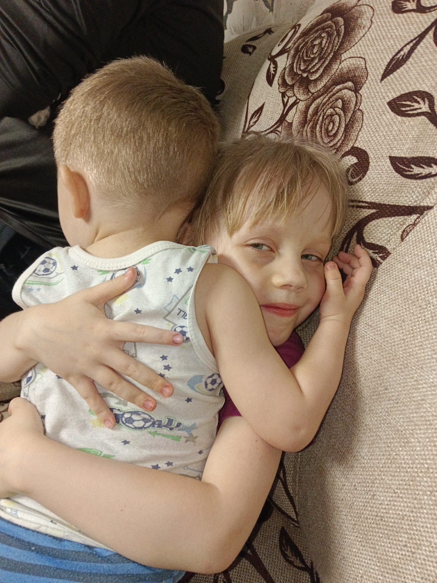Two young children are observed in an interior setting, identified as being in the city of Ольховка, Russia. A younger child, with short light-colored hair, is positioned with their back to the camera, wearing a white sleeveless undershirt patterned with blue stars and soccer balls, and blue and white striped lower garments. This child is embraced by a second, slightly older child, who has light-colored hair and light eyes, and is looking towards the camera with a subtle smile. The second child's left arm is wrapped around the first child's lower back, and their right hand is near their own face. The children are situated on a light-beige, textured fabric surface, likely a sofa or bed, which features a repeating dark brown floral pattern of roses and leaves. A dark, possibly black, fabric object is visible behind the first child.