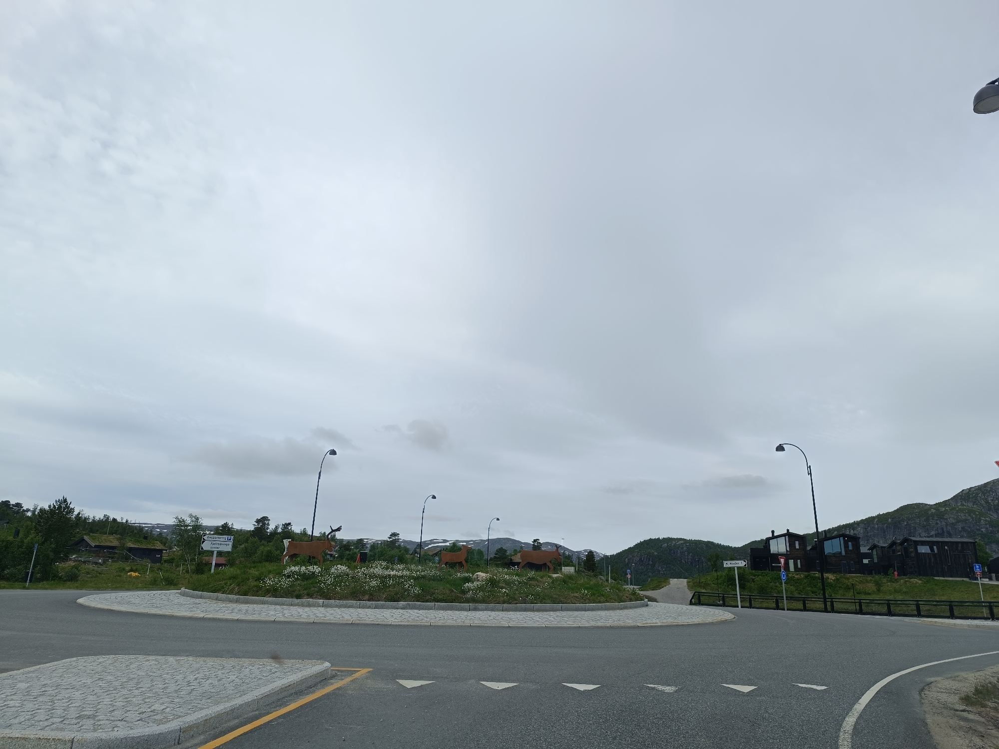 The image depicts a roundabout in Hovden, Norway, on a cloudy day. The roundabout features a circular grassy island decorated with white wildflowers and several large, cut-out silhouettes of reindeer. Three lampposts with curved arms are strategically placed around the roundabout, with one near the center island and two further out on the road.
In the background, to the left, there is a traditional Norwegian cabin with a grass roof. A white directional sign is visible on the left side of the roundabout, indicating "Østgardsvegen" and "Fjellgrenda/Hovden".
To the right of the roundabout, a modern black wooden building with large windows is situated on a hill. Further up the hill, dark green, steep forested slopes rise, some with patches of snow visible on the higher peaks, suggesting a mountainous environment. The road surface is asphalt with white triangular markings indicating traffic flow. A light-colored paved area with cobblestone-like texture is present on the left side of the foreground, bordered by a yellow line. The sky is overcast with light gray clouds.