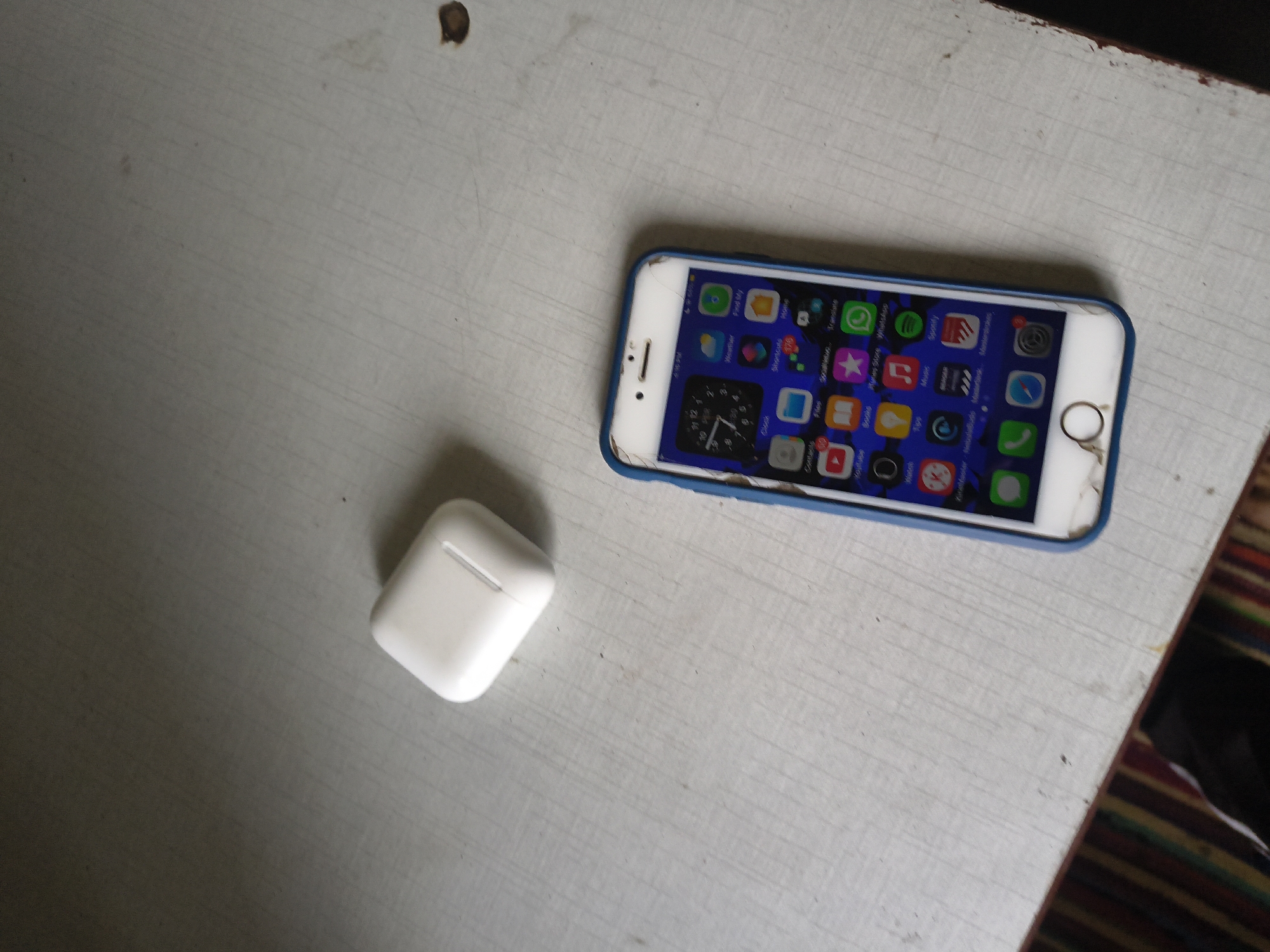 An iPhone 6s with a blue case and a white wireless earbud charging case are positioned on a light-colored, textured surface that appears to be a table. The iPhone's screen displays various app icons and a clock widget. The background is dimly lit and out of focus, showing a shadow and what looks like a striped pattern on the lower left. The scene is indoors.