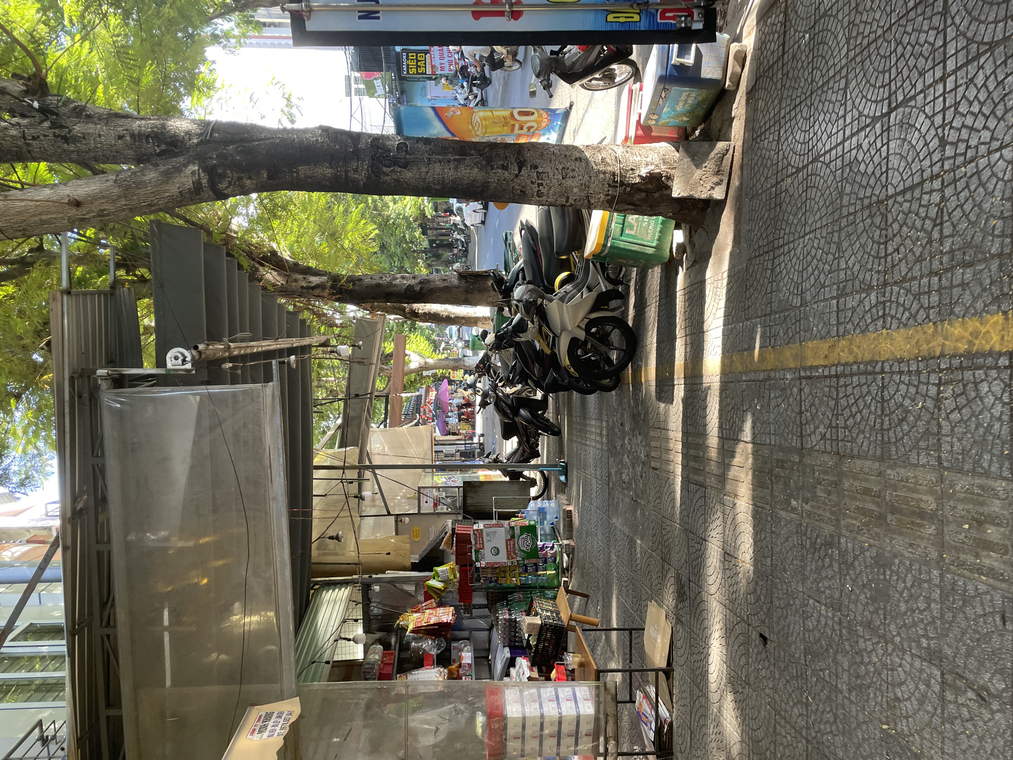 The image depicts a sunlit street scene in Da Nang, Vietnam. A sidewalk paved with patterned grey tiles runs parallel to a building with a covered walkway. The walkway houses a small shop with shelves stocked with various goods, including drinks and packaged items. Several motorcycles are parked along the sidewalk in front of the shop and further down the street. Trees line the sidewalk, casting shadows on the ground. A yellow line is painted on the sidewalk, indicating a traffic or pedestrian division. In the background, other buildings and a street with moving vehicles are visible, suggesting an urban environment. The lighting indicates it is daytime, likely late morning or early afternoon.