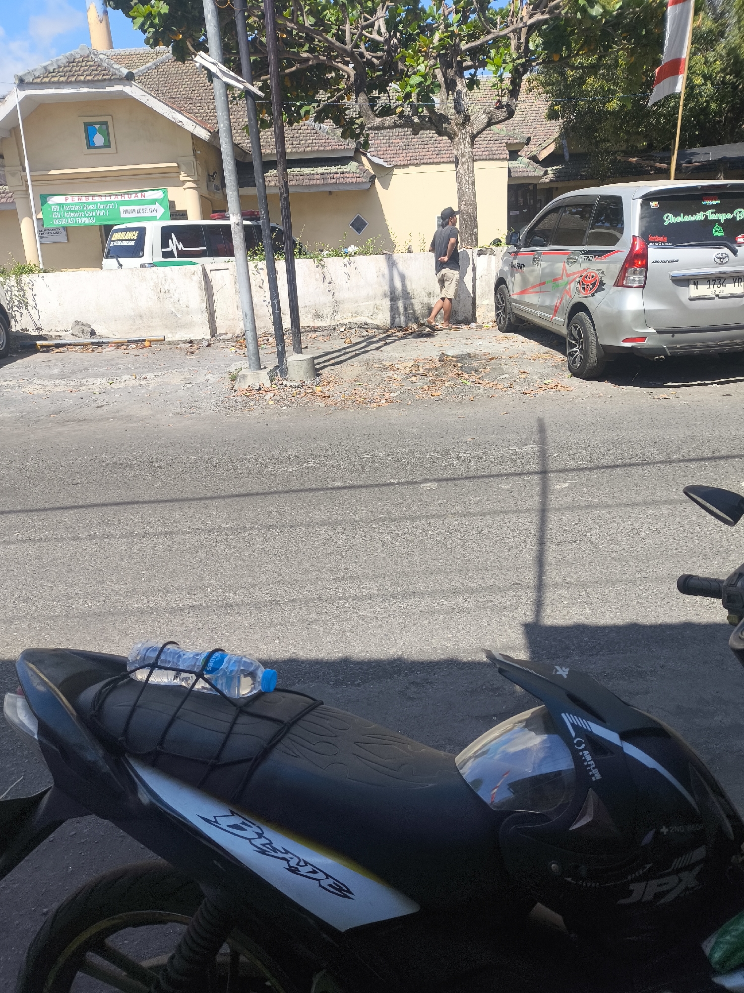 The image depicts a street scene in Lumajang, Indonesia. In the foreground, the back of a black motorcycle is visible, with a bottle of water secured to the seat by a black net. A black helmet with a visor is also placed on the motorcycle seat. In the background, a silver car with decals is parked on the side of the road. Adjacent to the car is a building with a yellow facade and a tiled roof. A banner with Indonesian text is displayed on the building. A white ambulance with green markings is parked in front of the building. A man in a black t-shirt and shorts is standing near the car, facing away from the camera. The environment appears to be a sunny day with scattered leaves on the ground.