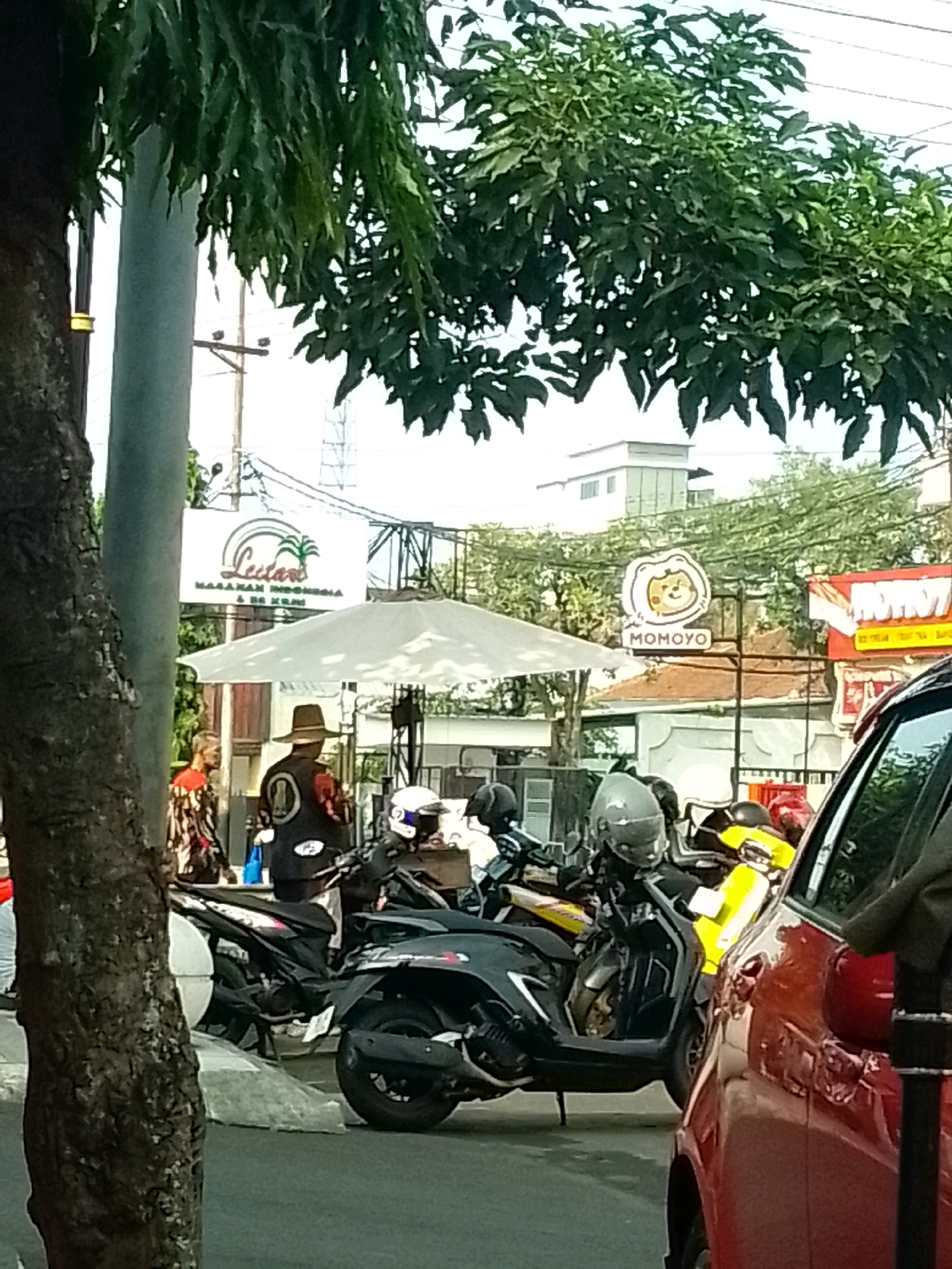 The image depicts a street scene in Jember, Indonesia, with a focus on parked motorcycles and a red car. In the foreground, a large tree trunk is on the left side. Behind it, a metal pole stands tall, partially obscured by foliage. A white canopy tent is set up, with several motorcycles parked underneath and around it. One man is visible wearing a patterned shirt and a hat, standing near a motorcycle. Another person is partially visible in the background.
Above the canopy, two signs are visible. The left sign features a palm tree logo and the text "Lutana Makanan Indonesia & Es Krim". To the right, a circular logo with a cartoon character and the text "MOMOYO" is displayed. Further to the right, a red and white sign with text is partially visible.
The environment appears to be a commercial or public area, possibly a street lined with shops or food vendors. Overhead wires and utility poles are present in the background, indicating an urban setting. The lighting suggests it is daytime. The red car on the right side of the image partially obstructs the view of the motorcycles and the surrounding area.