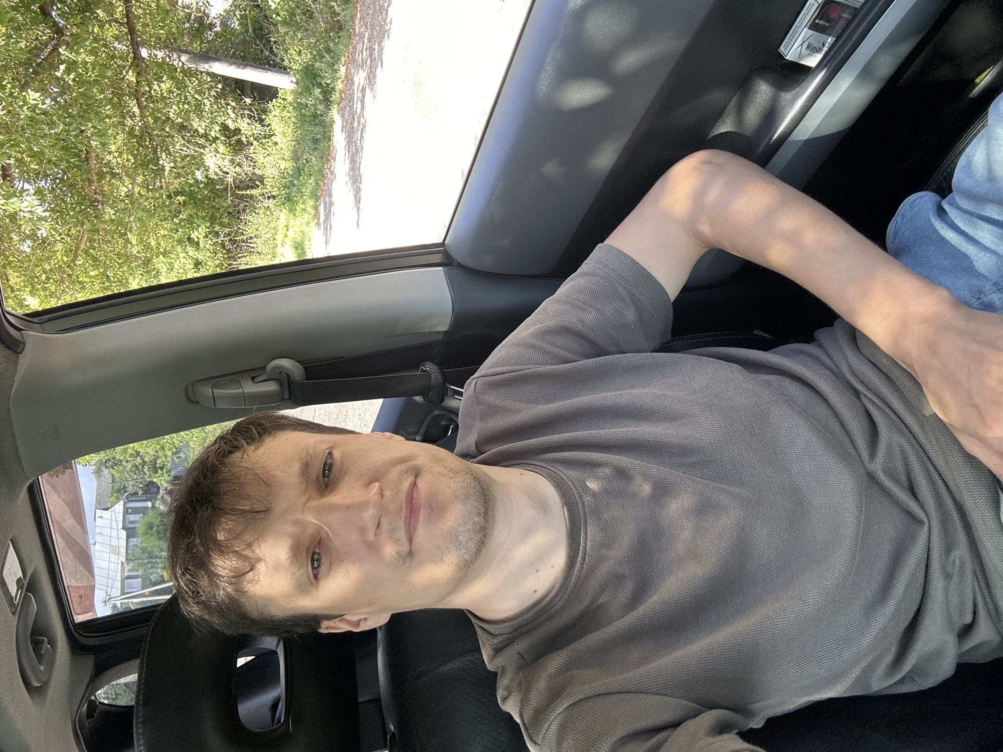 A selfie of a man taken from inside a car. The man has short brown hair, a light complexion, and appears to be in his late 20s or early 30s. He is wearing a grey t-shirt and blue jeans. His left arm is resting on the car door. The background shows a rural setting with trees and a dirt road. The location is the city of Makiv, Ukraine.