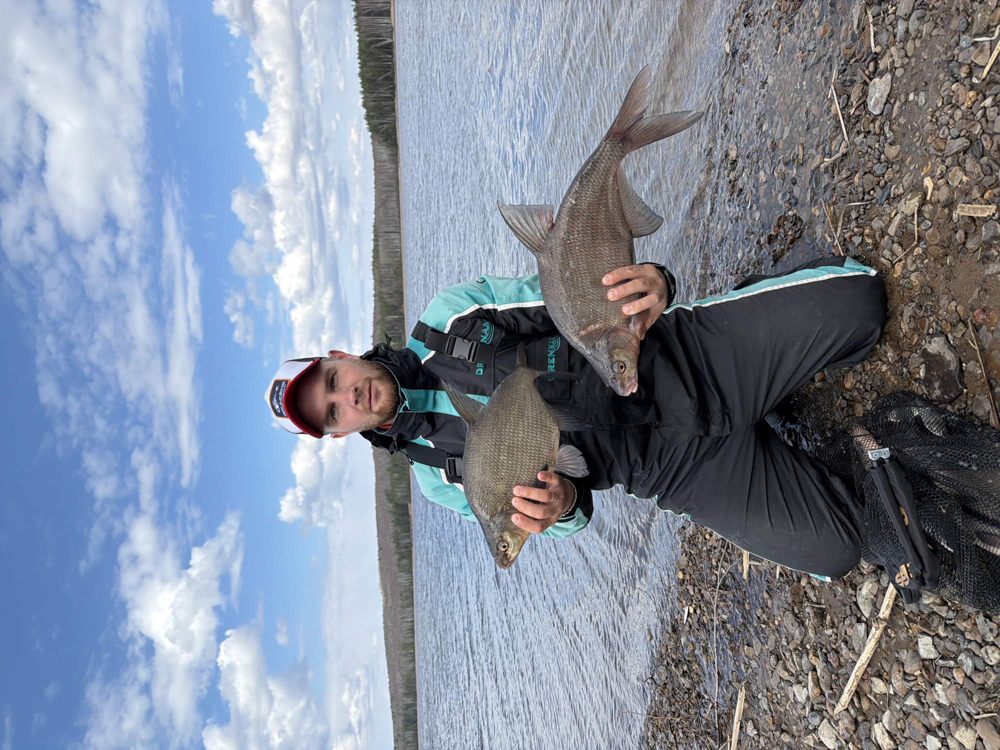 A man is depicted kneeling or squatting on a rocky shoreline next to a body of water, posing with two fish he has caught. The image is taken outdoors during daylight hours under a bright sky with scattered white clouds. The setting appears to be a lake or a large river, consistent with the Chaikovsky, Russia context, with a distant treeline visible across the water.
The man, with a beard and a neutral expression, wears a red and white cap, a black and teal fishing jacket or dry suit, and black pants. Over his jacket, he wears a black fishing vest or life vest. He holds two silvery-grey fish, likely bream, with both hands; one is significantly larger than the other. To his right, a black fishing net is visible on the ground.
Visible text includes "DFISH" on his jacket and "RENNA" partially visible on another section of his jacket. The black bag of the fishing net displays the brand name "SALMO" in white with red accents. The shore is composed of small, wet pebbles and some scattered debris. The water is relatively calm with small ripples reflecting the daylight.