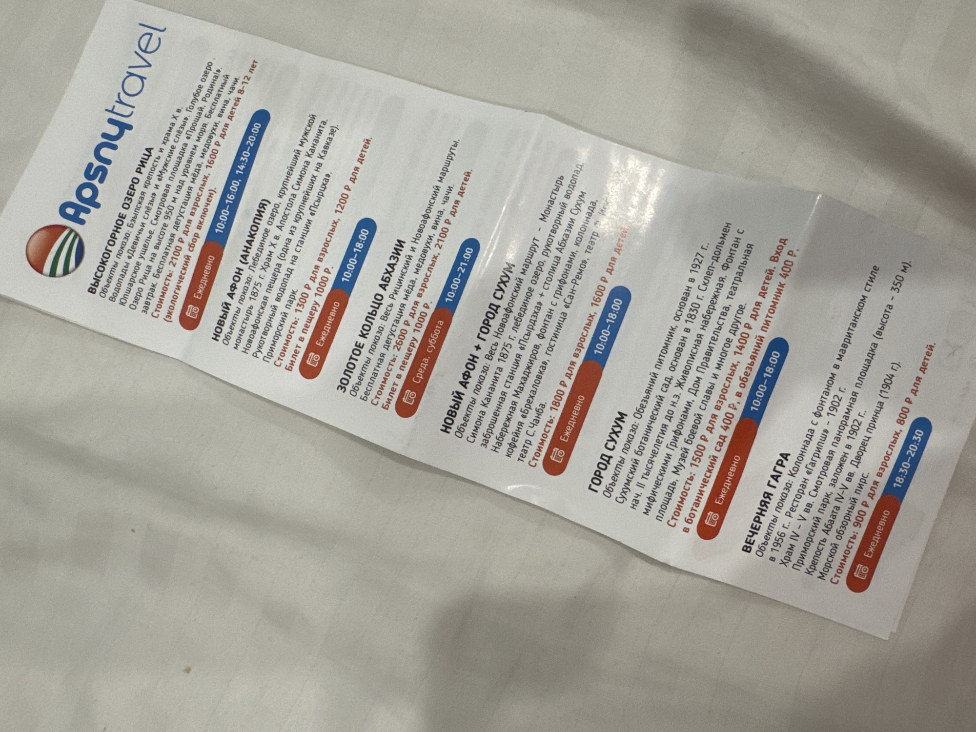 The image is a close-up of a printed brochure or flyer, likely an itinerary or advertisement for tourist attractions. The text is in Russian. The top of the flyer features a logo with a red, orange, and green semicircular design and the text "Apsnytravel". The content is organized into sections detailing different excursions or tours, each with a title, description of sights, and pricing.
Key sections include:
* **ВЫСОКОГОРНОЕ ОЗЕРО РИЦА (High-Mountain Lake Ritsa)**: Describes the lake, a viewing platform, a wine tasting, and a tour of the spa. It lists a price for adults and children.
* **НОВЫЙ АФОН (АНАКОПИЯ) (New Athos (Anakopia))**: Details the New Athos Monastery, the Simon Kananita Apostolic Church, a cave monastery, a hand-made waterfall, and Primorsky Park. Pricing for adults and children is provided.
* **ЗОЛОТОЕ КОЛЬЦО АБХАЗИИ (Golden Ring of Abkhazia)**: Lists sights such as Ritsa and Novoafonsky routes, a tasting of honey and wine, and a tour of various locations. Pricing for adults and children is included.
* **НОВЫЙ АФОН + ГОРОД СУХУМ (New Athos + City of Sukhumi)**: Describes a route including the New Athos Monastery, Lake Ritsa, a hand-made waterfall, the city of Sukhumi, the "San-Remos" fountain, a theater, and other amenities. Pricing for adults and children is specified.
* **ГОРОД СУХУМ (City of Sukhumi)**: Highlights the Sukhumi Botanical Garden, the Animal House, an ancient dolmen, the Government House, a fountain, the theater, and other attractions. It also mentions an entrance fee for the botanical garden.
* **ВЕЧЕРНЯЯ ГАГРА (Evening Gagra)**: Details sights such as the "Gagripsh" restaurant, a viewing platform, Primorsky Park, the Abkhazia IV-V century fortress, and the Palace of Prince A. K. Shervashidze, and the observation deck. Pricing for adults and children is listed.
Each section includes information about daily operation and specific time slots or hours. The overall aesthetic is clean and organized, with red and blue color accents used for emphasis.