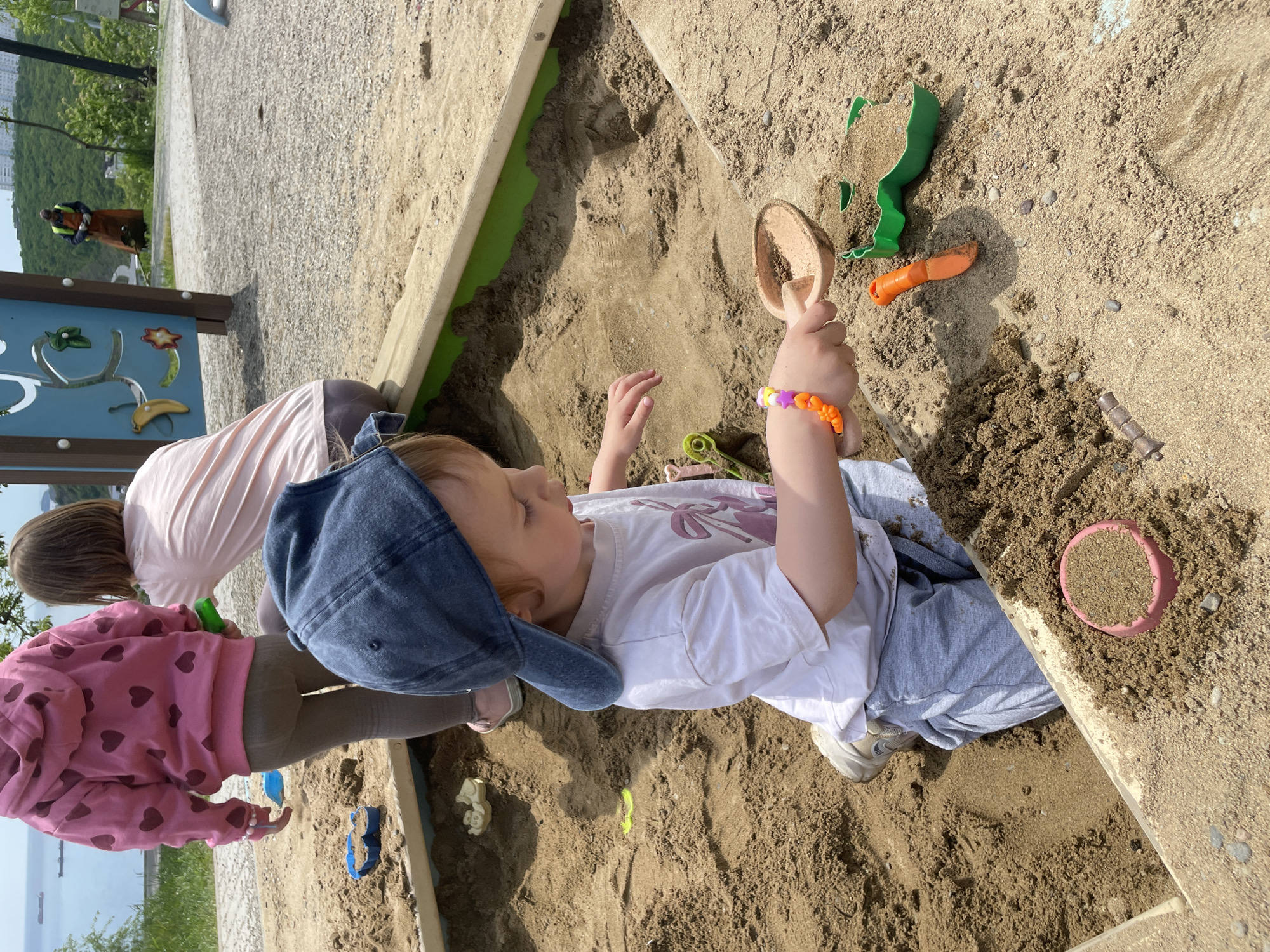 Two children are playing in a sandbox on a sunny day. One child, wearing a white t-shirt, grey sweatpants, a blue baseball cap, and a colorful bracelet, is holding a sand scoop above the sandbox. The other child, wearing a pink heart-patterned hooded sweatshirt and leggings, is playing near the edge of the sandbox. The sandbox contains various toys including shovels, a carrot-shaped toy, and a pink bucket. The sandbox is surrounded by wooden planks, with a green interior trim. In the background, there is a play structure and greenery. In the far background, a body of water and a cityscape can be seen. The scene appears to be in a park in Vladivostok, Russia.
