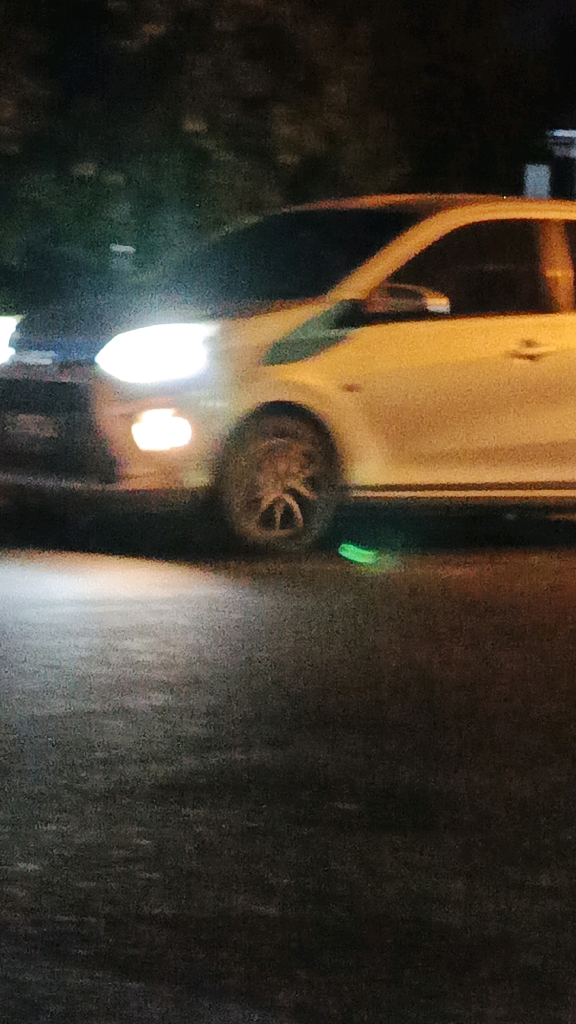 A slightly blurry, low-light image shows a portion of a white car in Surakarta, Indonesia. The car is on a dark, paved surface, possibly a road or parking lot, with trees visible in the background. The car's headlights are illuminated, casting a bright light. The front of the car is on the left side of the image, and the front passenger wheel is visible. A small, bright green object, possibly litter or debris, is on the ground near the front passenger tire. The overall impression is of a vehicle at night, possibly on a street or in a dimly lit area.