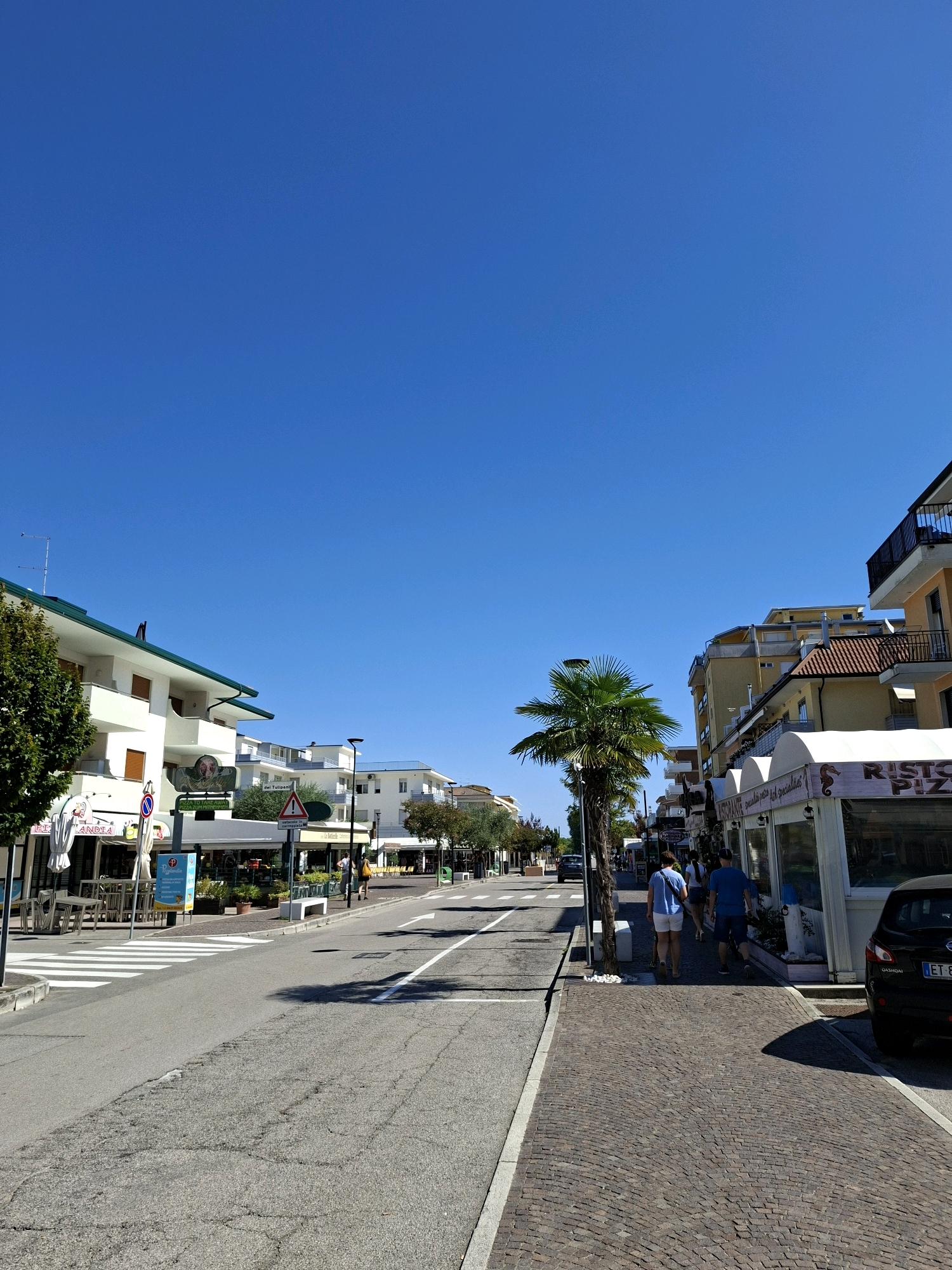 A street scene in Eraclea, Italy, on a clear, sunny day. The foreground shows a paved street with a crosswalk. To the right, a brick sidewalk is lined with shops and restaurants. A palm tree stands on the sidewalk. Two people, a woman in a blue shirt and white shorts and a man in a blue shirt and dark shorts, are walking away from the camera on the sidewalk. A black SUV is parked to the right of the frame. Buildings line both sides of the street, with balconies and awnings visible. The sky is a clear, vibrant blue.
