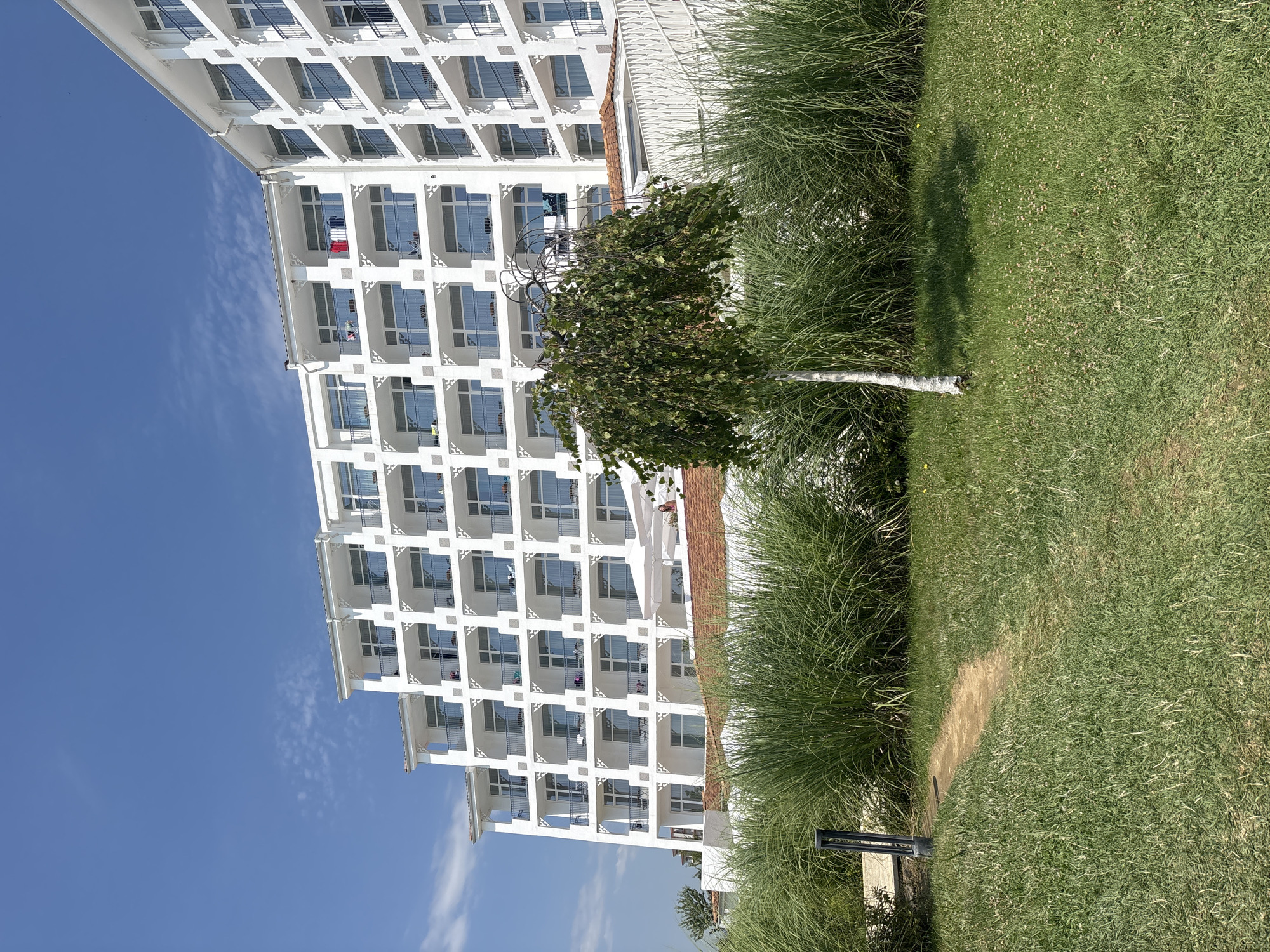 This is a photograph of a modern, multi-story white hotel building situated in the city of Byala, Bulgaria. The building has numerous balconies with black railings, and many windows. The hotel is set against a clear blue sky. In the foreground, there is a well-maintained lawn with a dirt path winding through it. Tall, green grass and a small tree with cascading branches are also visible in the foreground. A dark, modern lamp post is positioned to the left of the path. Beyond the hotel, some red-tiled roofs and a white fence are visible. There are no people clearly visible or interacting in the scene. The overall impression is one of a resort or vacation setting on a bright, sunny day.