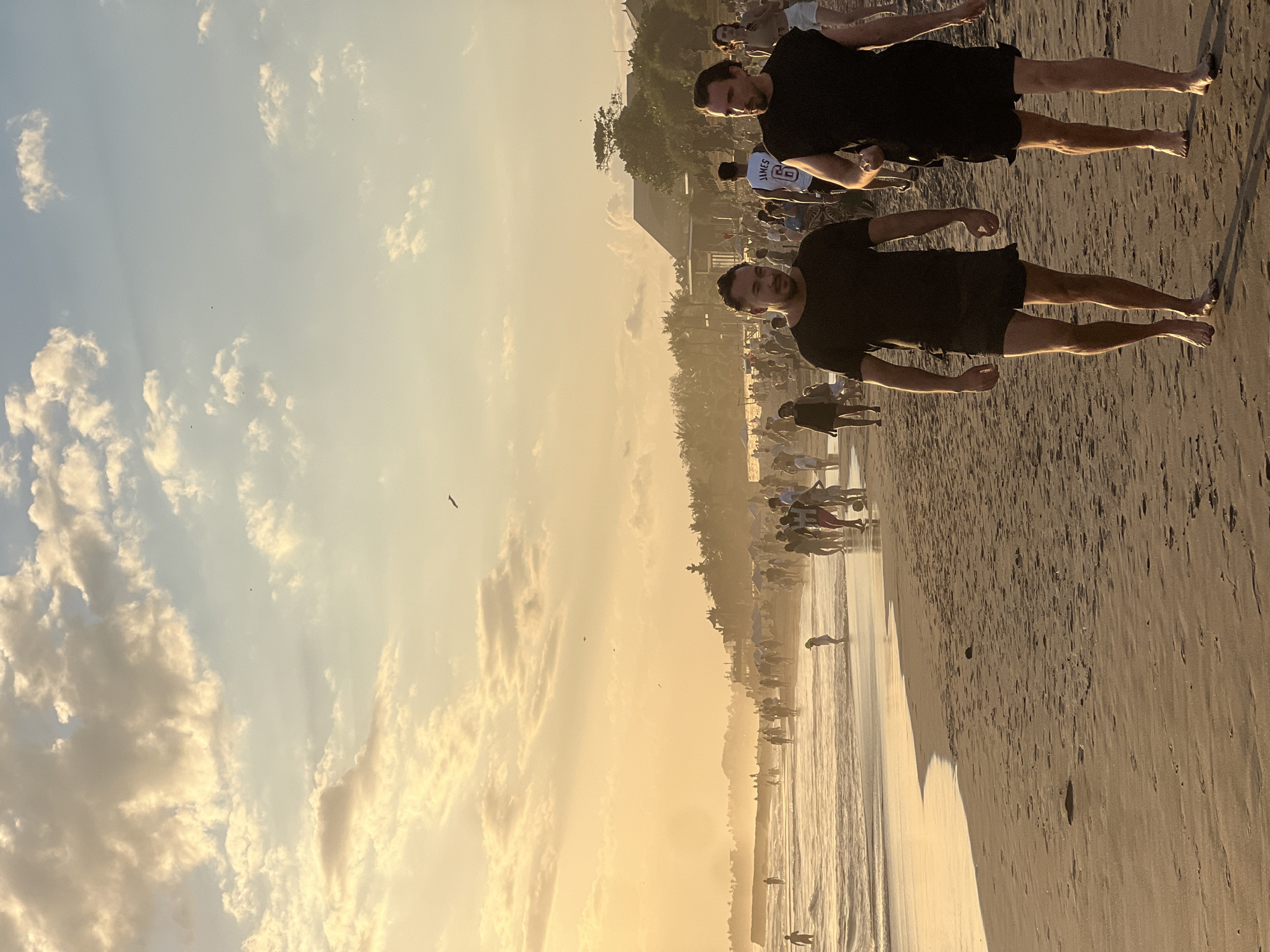 This image depicts a bustling beach scene in North Kuta, Indonesia, during sunset. The sky is illuminated with a warm, golden hue, casting a glow over the sand and water. Numerous people are present, walking along the shoreline, playing in the shallow waves, or gathered in groups. In the foreground, two men are walking towards the viewer, dressed casually in dark t-shirts and shorts. The beach is sandy with visible footprints and some scattered debris. The ocean has gentle waves breaking on the shore, and further back, a line of trees and buildings can be seen, suggesting a developed coastal area. The overall atmosphere is one of leisure and activity.