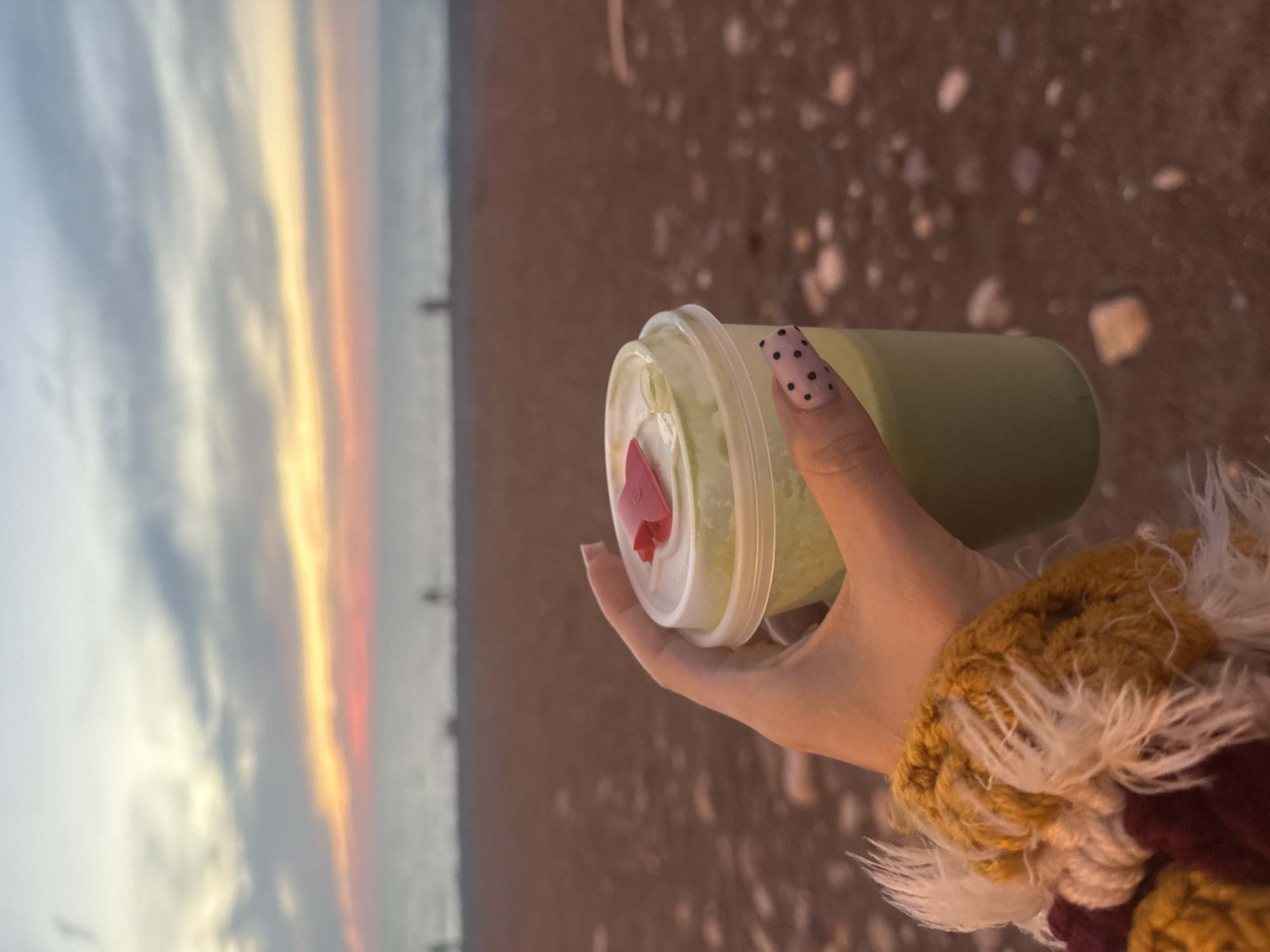 A photograph captures a person's hand holding a green beverage in a clear plastic cup against a blurred beach scene. The hand is adorned with black polka-dot nail art on one nail, and the person is wearing a colorful knitted garment with white, mustard yellow, and maroon threads. The cup features a clear lid with a red heart-shaped straw stopper. The background depicts a beach with a sandy or pebbly surface. In the distance, the sky displays hues of orange, yellow, and gray, suggestive of a sunset. The image is slightly blurred and seems to be taken in Batumi, Georgia, based on the beach and sunset colors which match known imagery of that location.