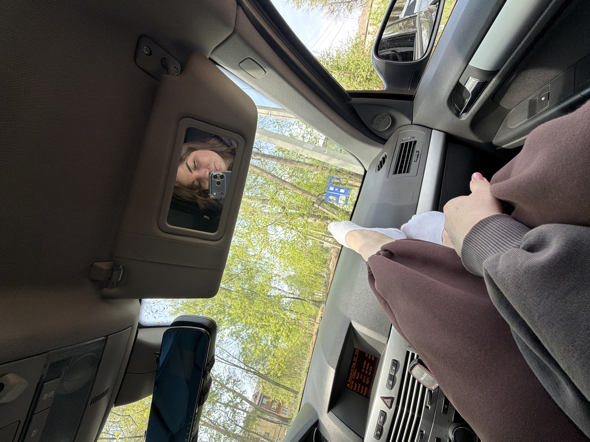 The image, taken from inside a car, shows a woman's legs on the dashboard. She is wearing brown pants and white socks. The car's dashboard features a climate control display showing "19.0°C" and other parameters, as well as a "WALKER" brand object near the climate controls. In the rearview mirror, the driver's face, partially obscured by a phone, is visible. Outside, trees suggest a park or residential area. The location in the city of Chaikovsky, Russia, cannot be confirmed from this image alone.