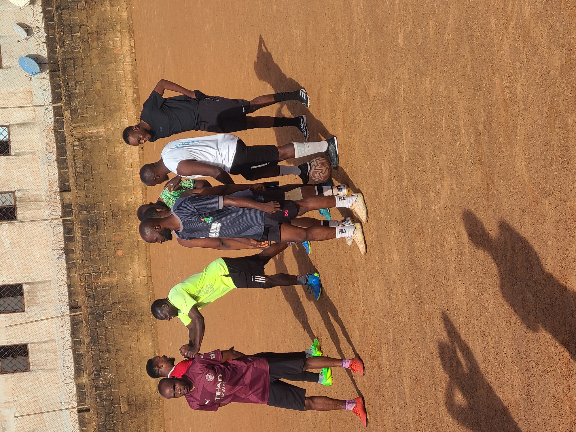 A group of seven men are standing on a dirt field in what appears to be Barakin Lerawi, Nigeria. They are dressed in athletic attire, including t-shirts, shorts, and cleats. Some of the men are interacting with each other, with two of them shaking hands. A soccer ball is visible on the ground between two of the men. In the background, there is a brick wall topped with barbed wire, and a building with barred windows. The sky is a clear blue. The scene appears to be a casual gathering or practice session on a sports field.