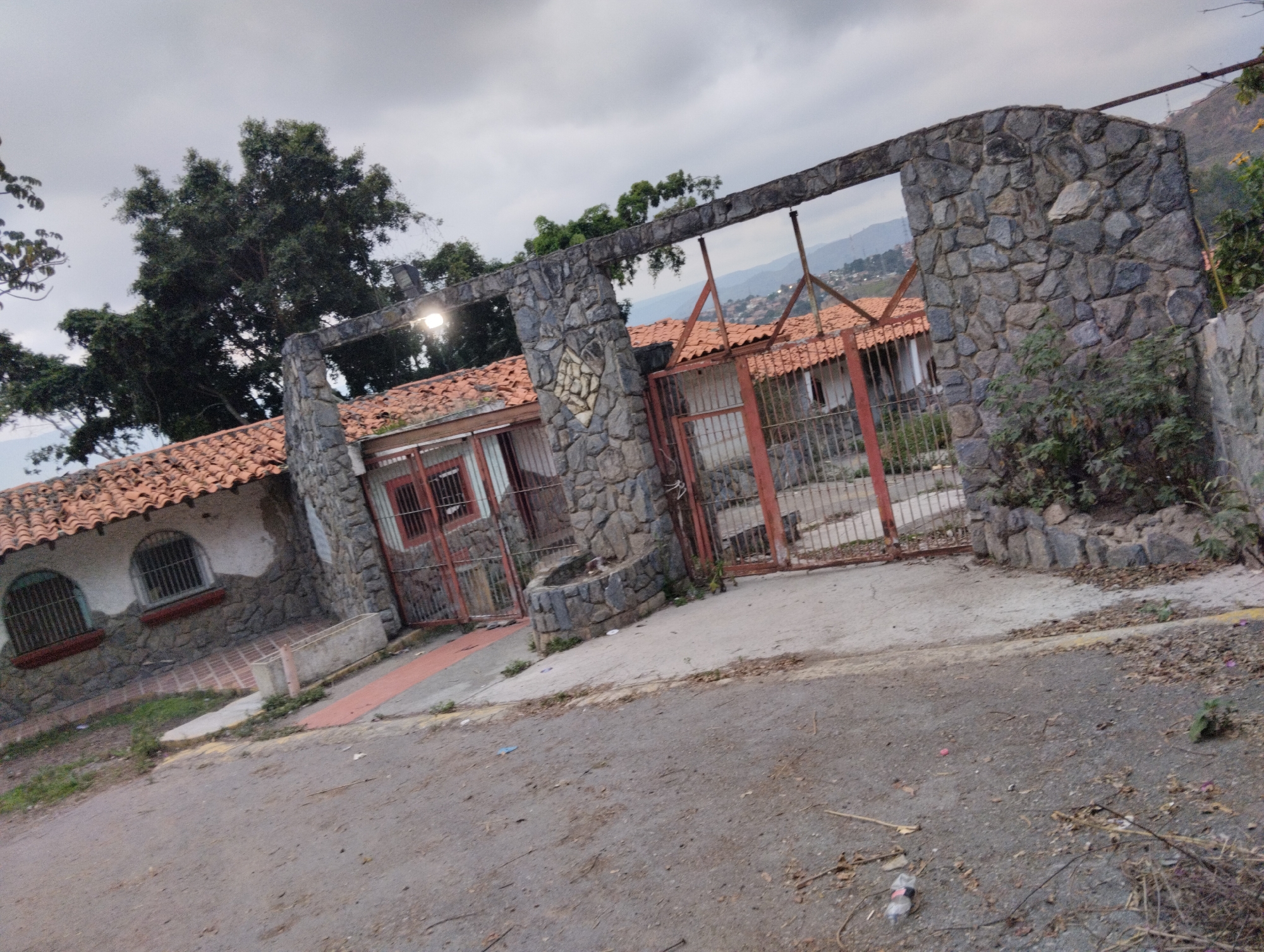 The image depicts the entrance to a property in what appears to be a hillside or mountainous area. The entrance features a stone archway with rusty metal gates. The property behind the gates has buildings with red tile roofs and stone or masonry walls. The scene is somewhat overgrown, with vegetation visible around the entrance and on the property. The road leading up to the entrance is paved but shows signs of wear and tear. The sky is overcast, suggesting a cloudy day. The location may be Caracas, Venezuela, based on the architecture and mountainous terrain, but this cannot be verified without more information.
