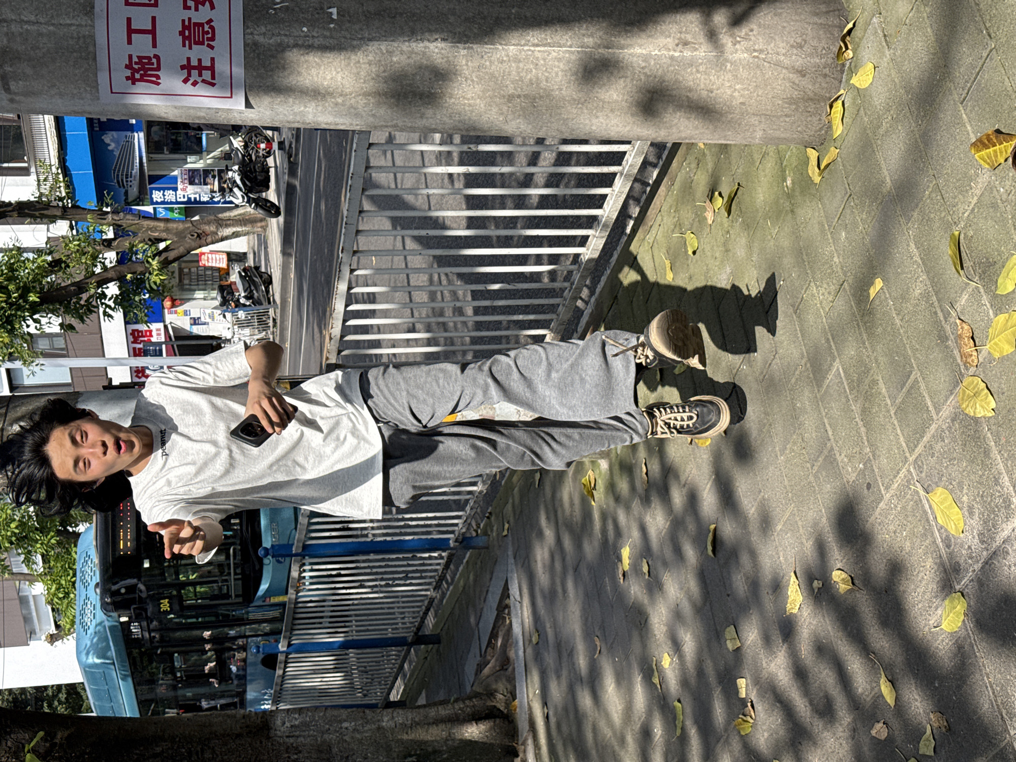 A young man of East Asian descent stands casually on a sunlit sidewalk in an urban setting, likely Nan'an District, China. He is dressed in a light grey or white t-shirt with "peanut" written in small letters on the chest, paired with grey sweatpants and black and white sneakers. He holds a black smartphone in his left hand and extends his right hand with an open palm or pointing gesture. His face shows an open-mouthed expression with his eyes slightly squinted, possibly reacting to the bright sunlight or posing playfully for a photo.
Behind the man, a blue public bus with the route number "304" and Chinese characters is partially visible. Further back, parked motorcycles can be seen. To the man's left, a sturdy concrete pole rises, displaying a rectangular sign with red borders. The sign contains Chinese characters that read "注意施工" (Zhùyì shīgōng), translating to "Attention: Construction" or "Notice: Construction." A metal railing or fence runs along the sidewalk, separating it from the street.
The sidewalk itself is paved with light grey blocks and is covered with scattered yellow leaves, suggesting an autumn season. Strong, clear shadows from overhead trees and the pole stretch across the pavement, indicating a bright, sunny daytime. In the background, buildings and green foliage complete the busy street scene.