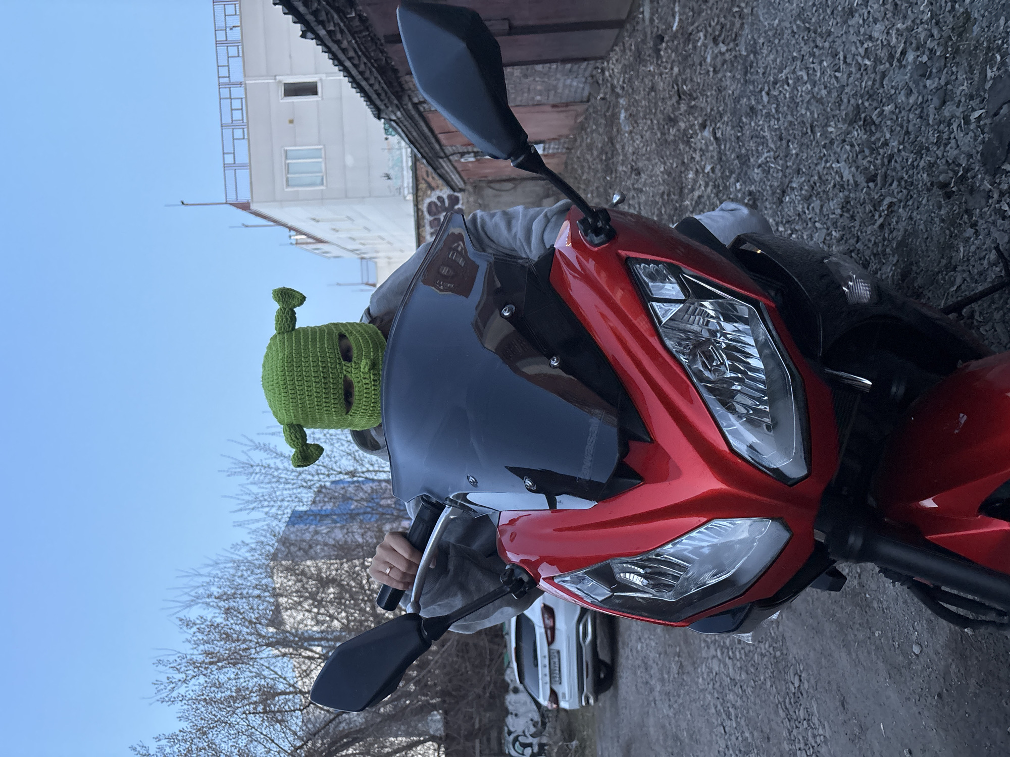A person wearing a green Shrek-themed balaclava is sitting on a red motorcycle. The motorcycle has black rearview mirrors and a black windshield. The person is holding the handlebars. The background includes a building, bare trees, a white car, and a gray paved surface. The location is Krasnoyarsk, Russia.