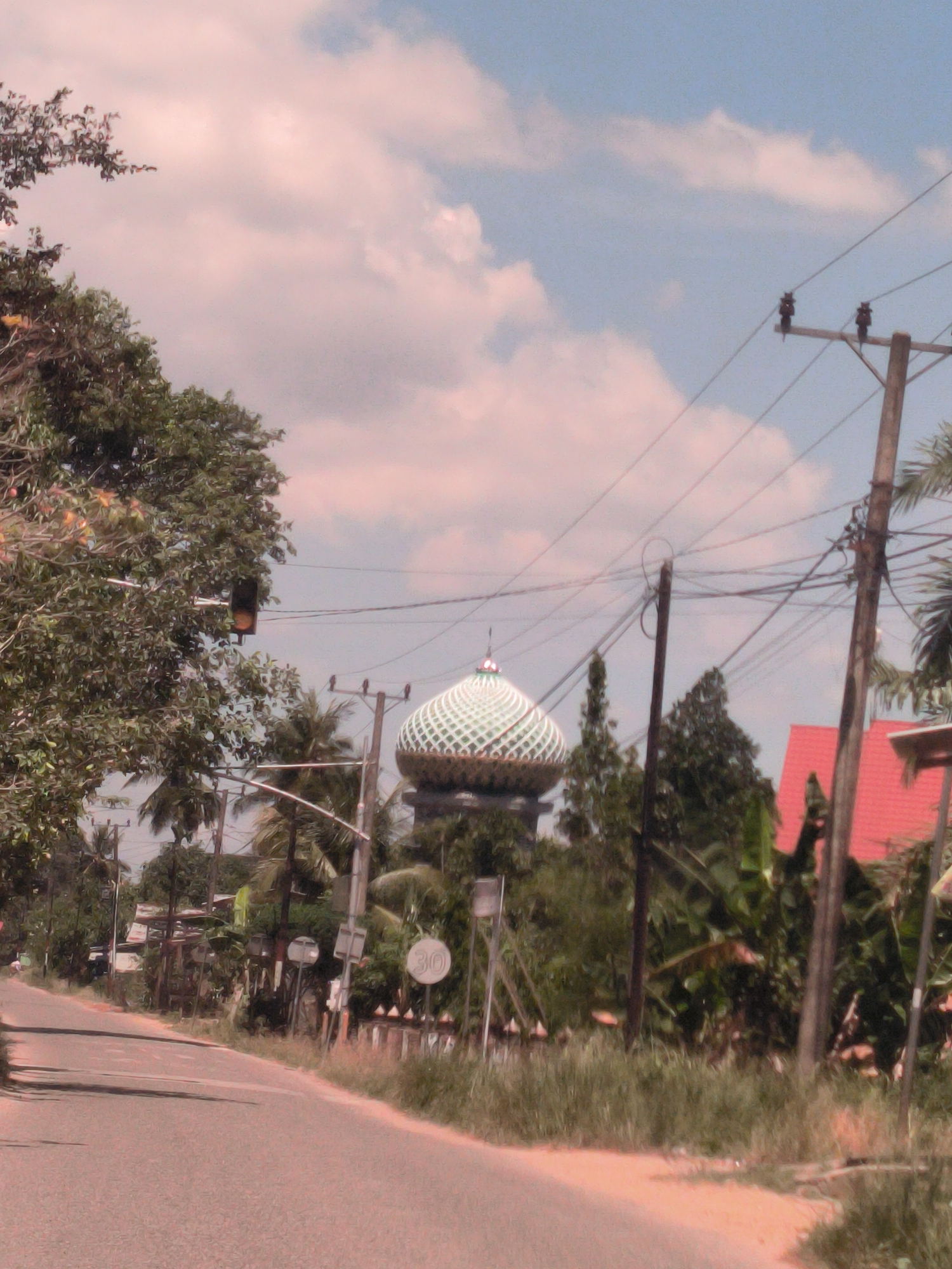 A street scene in Merbopuntong, Indonesia shows an asphalt road curving to the left, bordered by vegetation. Overhead power lines connect wooden utility poles. A mosque with a green and white patterned dome rises beyond the trees, with a traffic light visible near the road. A speed limit sign displays "30". A red roof is visible on a building to the right of the road. The sky is partly cloudy.