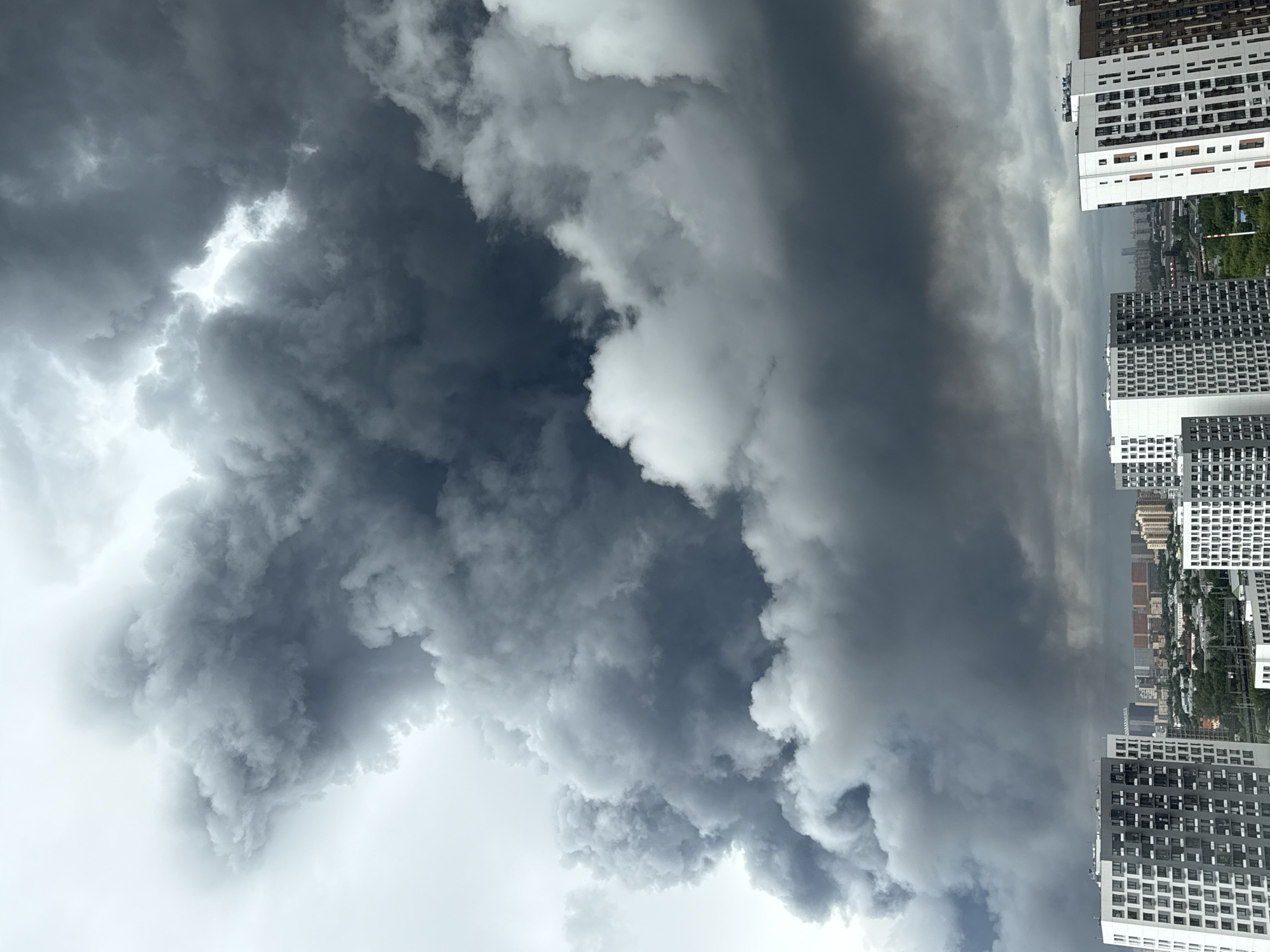 The image shows a large plume of dark smoke rising into a cloudy sky over the city of Tomilino, Russia. The smoke appears to be coming from a fire or industrial event. In the foreground, there are several modern apartment buildings. The sky is overcast and a bright light is visible through a gap in the clouds, suggesting the sun is trying to break through. The overall impression is one of an urban environment affected by a significant event, possibly an industrial accident or a large fire.