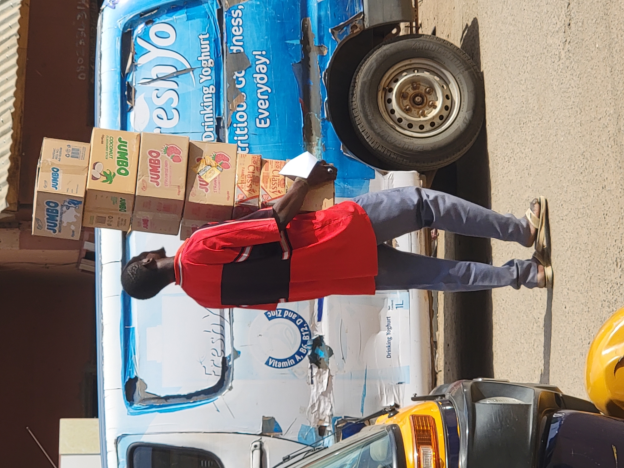 The image, captured in Maiburiji, Nigeria, depicts a man engaged in what appears to be a delivery or inventory activity on a bright, sunny day. The man, dressed in a red and black short-sleeved shirt, blue trousers, and sandals, stands next to a blue and white delivery vehicle, likely a van. He is focused on a stack of cardboard boxes resting on the side of the vehicle, holding a white piece of paper in his right hand, possibly a manifest or checklist.
The delivery vehicle features prominent branding. Its upper section is blue, while the lower part is white, and the paint shows some wear. A wheel with a silver hubcap is visible on the right side of the vehicle. In the foreground, parts of another vehicle, characterized by yellow and dark blue colors, are partially visible, possibly a tricycle or a small utility vehicle. The ground appears to be a paved or concrete surface, and strong shadows suggest a mid-day sun.
Numerous texts are visible on the vehicle and the boxes. On the side of the blue and white vehicle, text includes "Freshyo", "Drinking Yoghurt", "trition dness,", "Everyday!", "Vitamin A, B6 B12, D and Zinc", and "1L". The stacked cardboard boxes primarily feature the brand "JUMBO", with some boxes specifying flavors like "COCONUT FLAVOURED STICK" and "STRAWBERRY". Other visible text on the boxes includes "Classy", "PHASDAT", and "020".