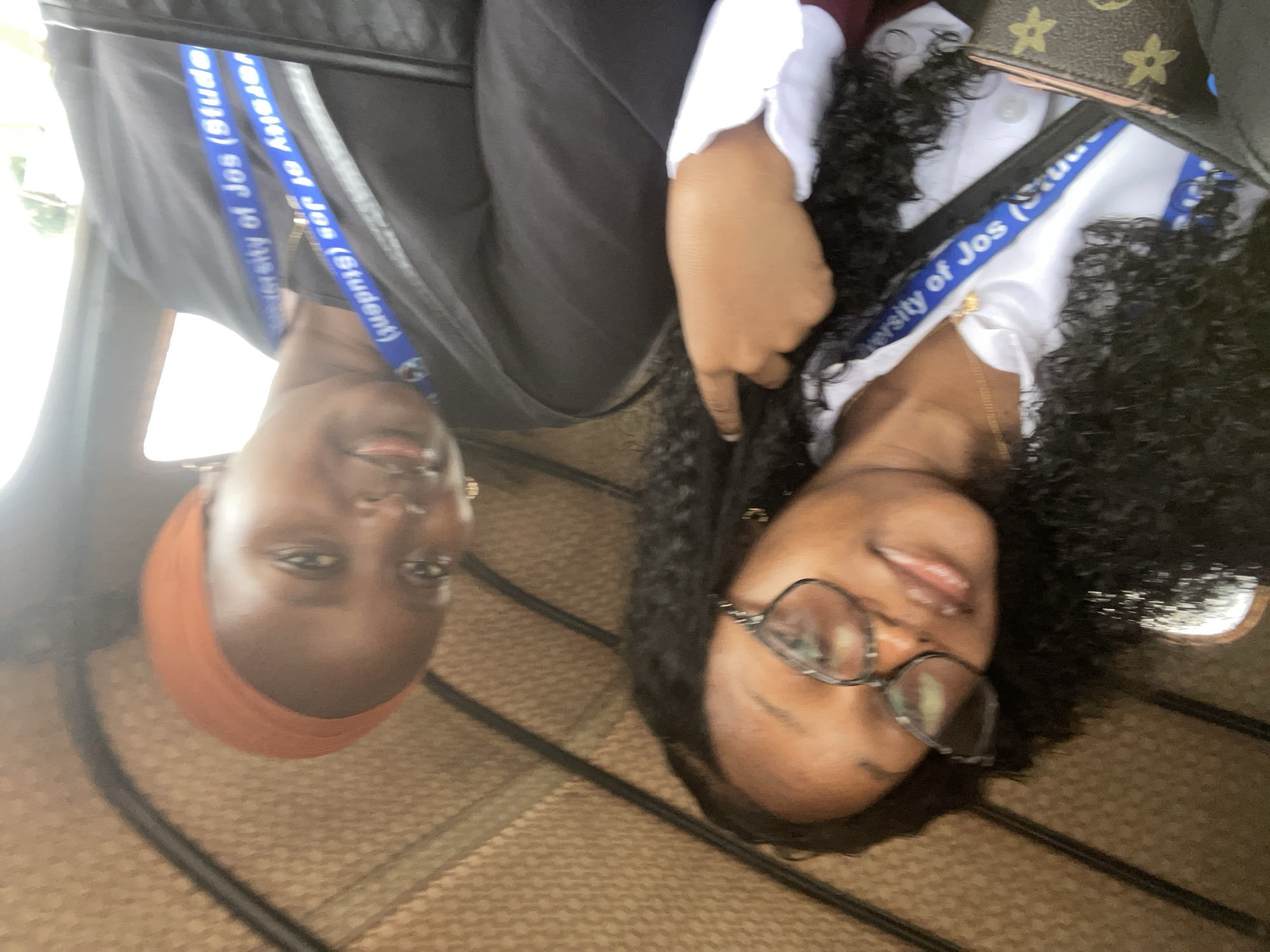 Two young women are inside a motorized rickshaw, likely in Rafin Par, Nigeria. The woman on the left has curly black hair and is wearing glasses and a white collared shirt. A blue lanyard with the text "University of Jos (Student)" is around her neck. She is holding a small, brown purse with a floral pattern. The woman on the right is wearing an orange headscarf and a black top. She also has a blue lanyard around her neck with the same "University of Jos (Student)" text. The interior of the rickshaw has a textured, woven ceiling and is brightly lit by natural light, suggesting it is daytime. The overall scene depicts two students on the go.