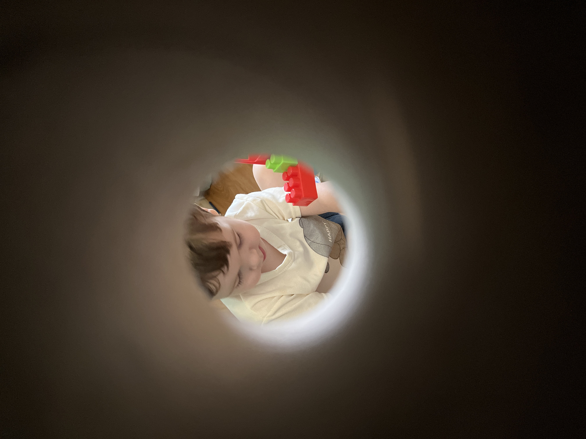 A child is seen through a circular opening, likely a tube or pipe, wearing a light-colored long-sleeved shirt with a bear design on the shoulder, playing with red and green building blocks. The background within the circle is a wooden floor. The location in Chaikovsky, Russia, is unknown from the image alone.