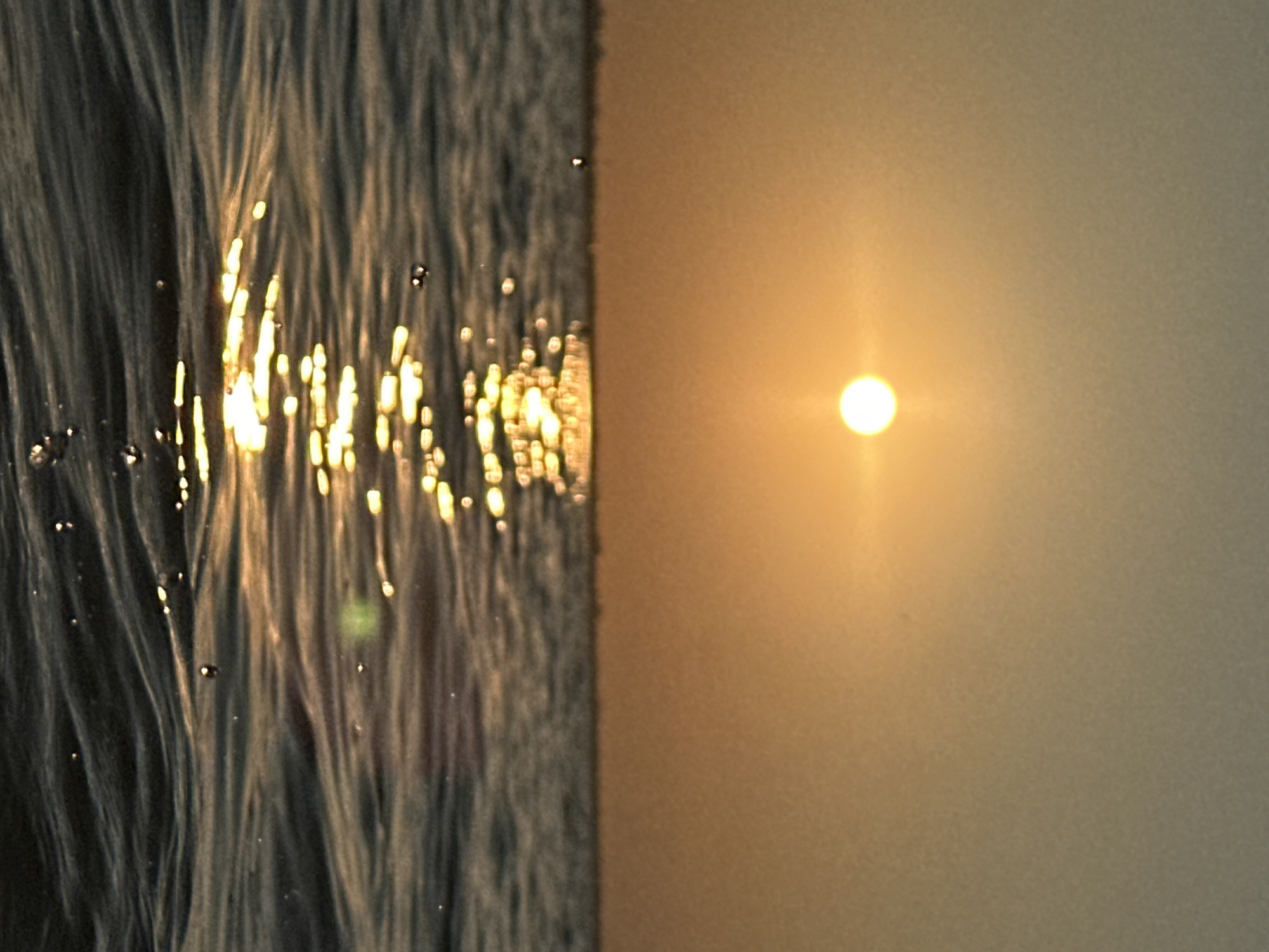 This is a close-up, low-angle shot of the sun setting over a body of water. The sun is a bright, diffused orb in the upper center of the frame, casting a shimmering reflection on the water's surface. The water itself is choppy, with small waves and ripples catching the golden light. There are a few water droplets visible in the foreground. The sky is a gradient of warm oranges and yellows, transitioning to a muted grey at the top of the frame. There are no people or man-made structures clearly visible in the image, though a faint, blurry line suggesting a distant horizon or landmass is present. The overall impression is one of natural beauty and tranquility. The image does not contain any elements that would indicate its location in Kokshetau, Kazakhstan.