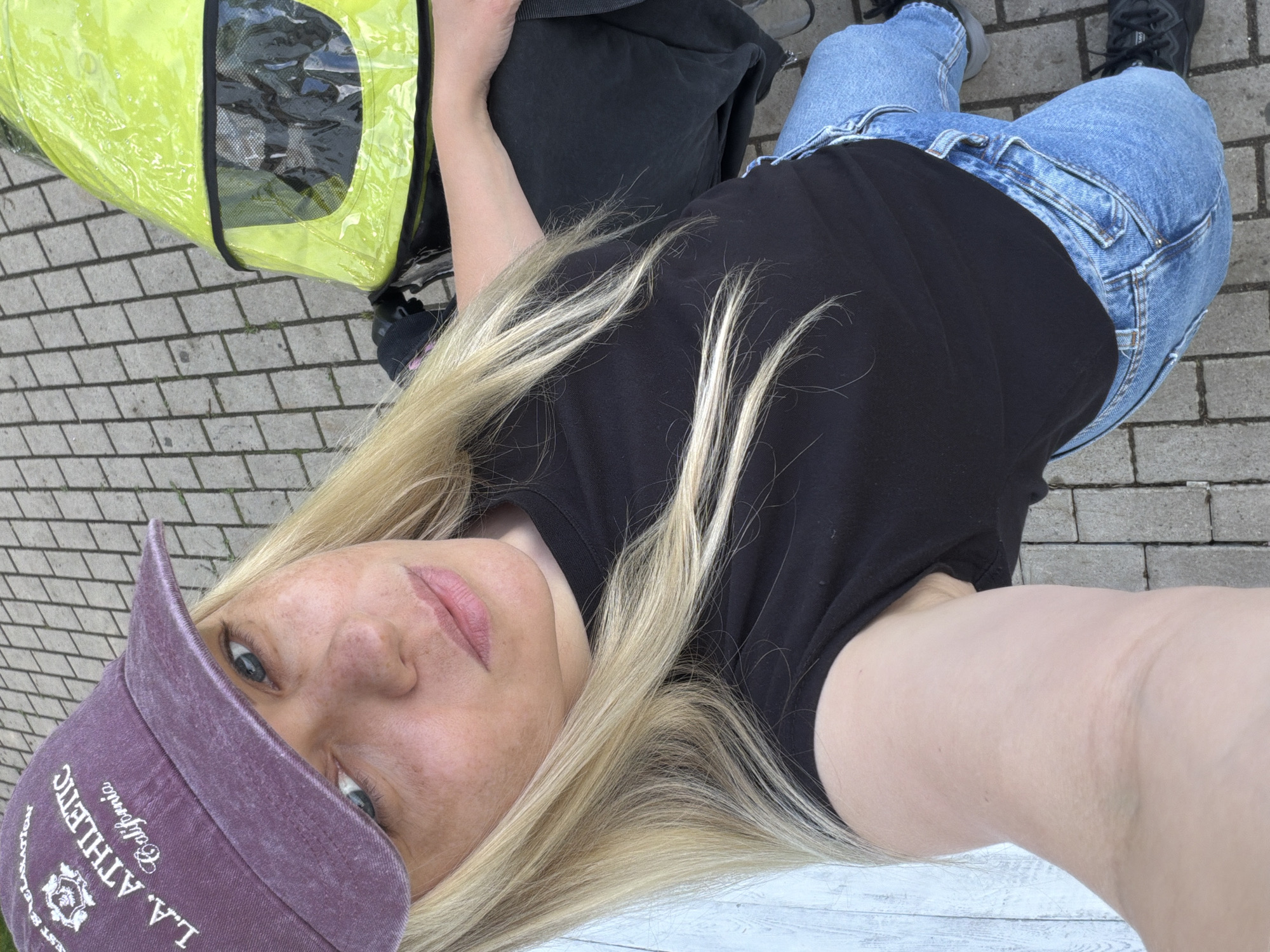 A top-down, selfie-style image shows a woman wearing a purple baseball cap, black t-shirt, and blue jeans, standing on a paved surface. A green and black wheeled carrier is positioned to the left of the woman. The photograph was likely taken outdoors, judging by the daylight. The exact location in Votkinsk, Russia is not discernible from the visual elements of the photograph.