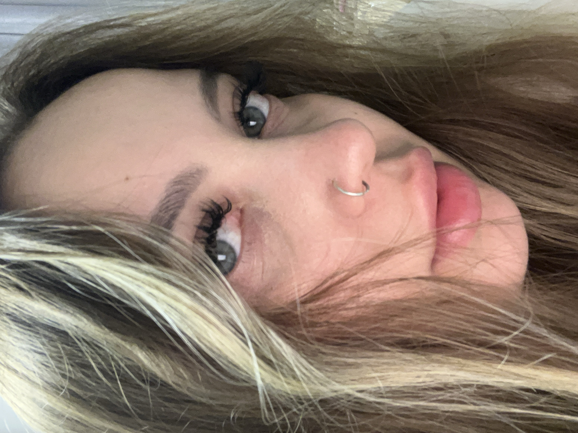 A close-up, slightly tilted image shows a woman's face. She has light brown and blonde hair framing her face, prominent eyelashes, and light-colored eyes. A silver ring is visible in her left nostril. Her lips are slightly parted and appear pink. The background is indistinct, suggesting the focus is entirely on her face. The image does not provide any information suggesting the location to be Pritsepilovskaya, Russia.