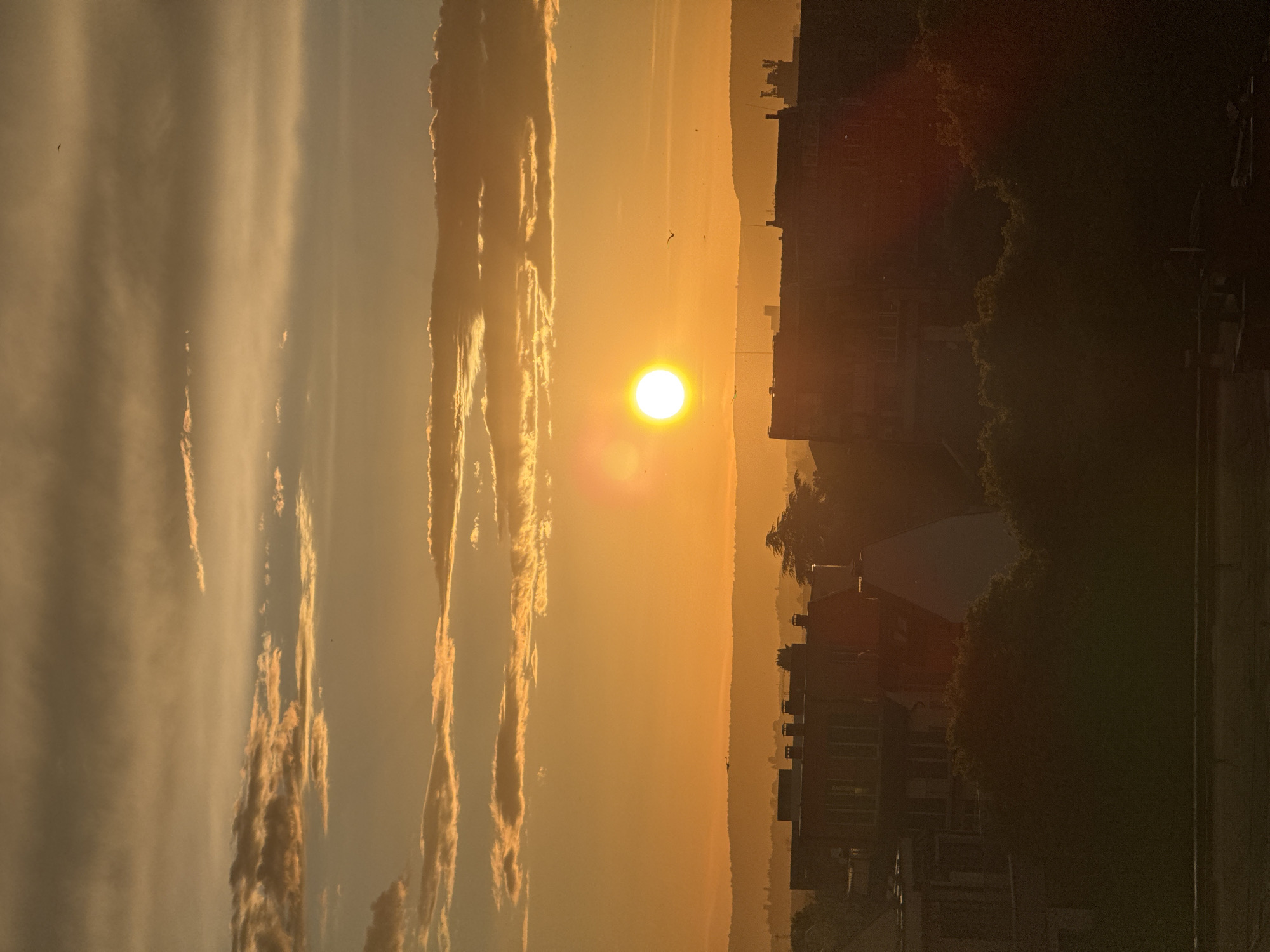 The image depicts a sunrise in Varna, Bulgaria. The sun is positioned centrally in the frame, emitting a bright orange glow. To the left, striated clouds are visible. To the right, a cityscape silhouette emerges, with buildings, trees, and infrastructure discernible. The overall scene conveys a sense of urban life juxtaposed with natural beauty.
