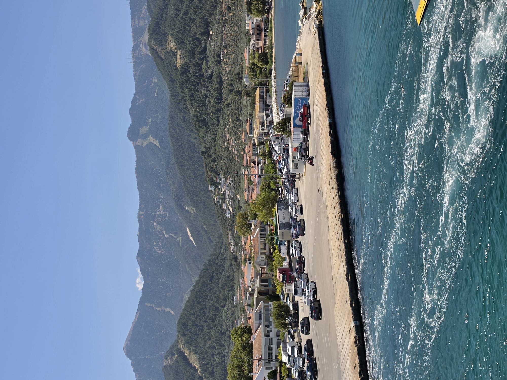 A wide, high-angle, full shot captures a sun-drenched coastal town on a clear, bright day. The foreground is dominated by the turquoise blue waters of a harbor, churned into white foam by a boat's wake. To the right, a bright yellow object is partially visible in the water.
The midground features a concrete pier extending into the water, laden with numerous parked cars of various colors, including black, white, silver, and red. A red truck is also parked on the pier, alongside several white shipping containers. A small, white structure, possibly a booth or small building, sits near the beginning of the pier, with a red motorcycle parked in front of it.
Beyond the pier, a coastal town unfolds, characterized by buildings with terracotta-tiled roofs and white or light-colored walls. Green trees are interspersed throughout the town, suggesting lush vegetation. The town is nestled at the base of a verdant, tree-covered mountain range that fills the background. The mountains are rugged, with visible ski slopes or trails ascending their sides, and several communication towers are scattered across the higher elevations. The sky above is a clear, vibrant blue.