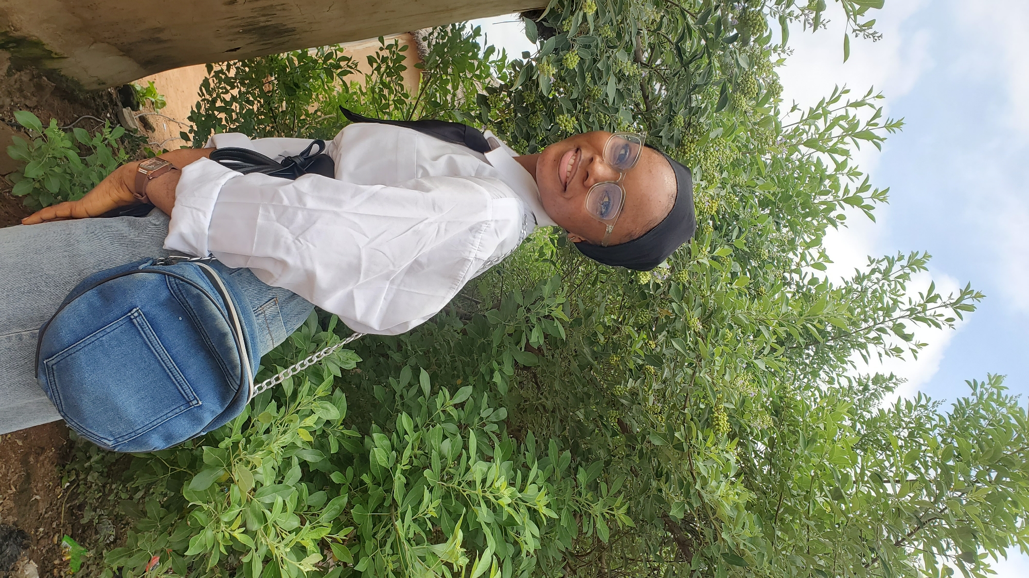 A young Black woman wearing a black hijab, clear-rimmed glasses, a white button-down shirt with a black tie, and light-wash denim jeans is standing outdoors in Jalingo, Nigeria. She is wearing a blue denim crossbody bag with a silver chain strap. She is smiling and looking to her left. The background consists of lush green foliage and a cloudy sky. A tree trunk is visible on the left side of the frame. The ground appears to be dirt.