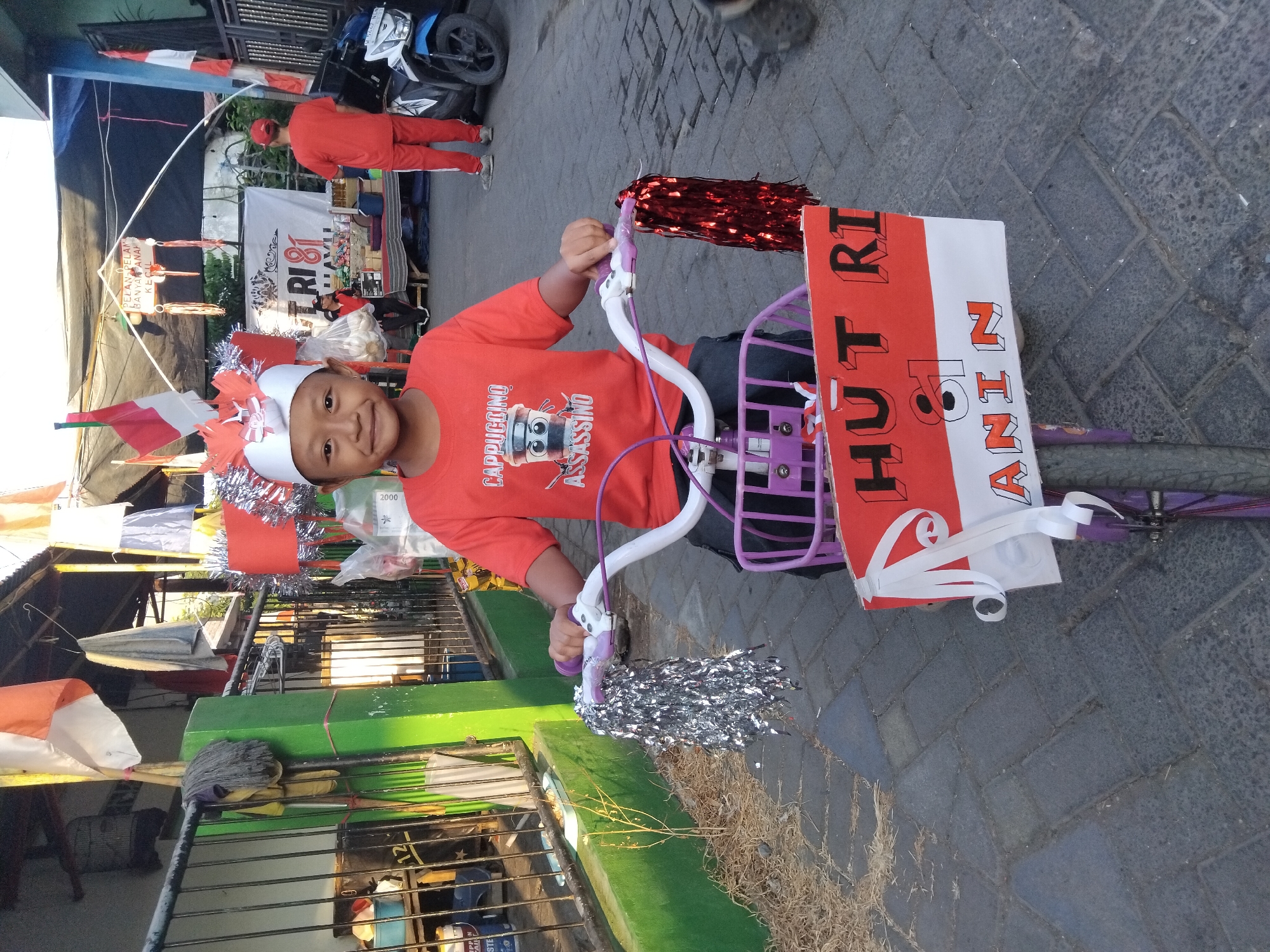 A young Indonesian boy, appearing to be between five and seven years old, sits on a purple bicycle with a basket. He wears a red t-shirt with a graphic of a cartoon coffee cup labeled "CAPPUCCINO ASSASSINO" and a white festive headpiece adorned with red and silver decorations. The bicycle is decorated with red and white streamers on the handlebars, a purple basket, and a sign on the front that reads "HUT RI 81" in red and black lettering on a red and white background, with the word "ANIN" below it in orange and white. He is outdoors, on a paved sidewalk in what appears to be a residential area. Behind him, a banner reads "HUT RI 81" and other decorations suggest a celebration, possibly for Indonesian Independence Day. In the background, two adults in red outfits are visible, one facing away from the camera and another attending a stall. A blue motorcycle is parked to the right. The scene is set in Kepuh Kiriman, Indonesia.