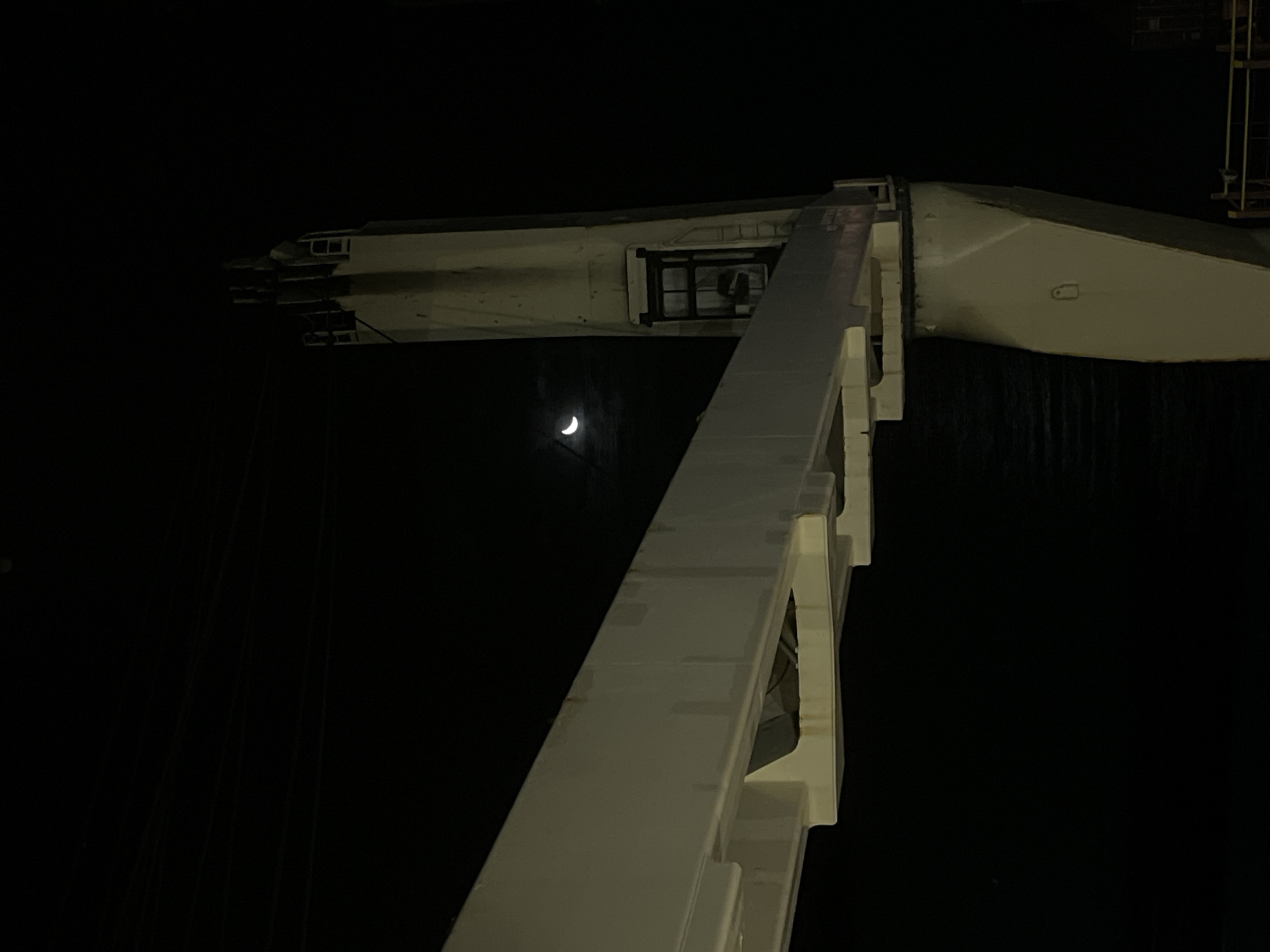 The image shows a low-angle view of a white crane boom at night. The moon is visible in the background between the crane boom and the arm of the crane. The scene is dark, with the crane and moon being the primary sources of light.