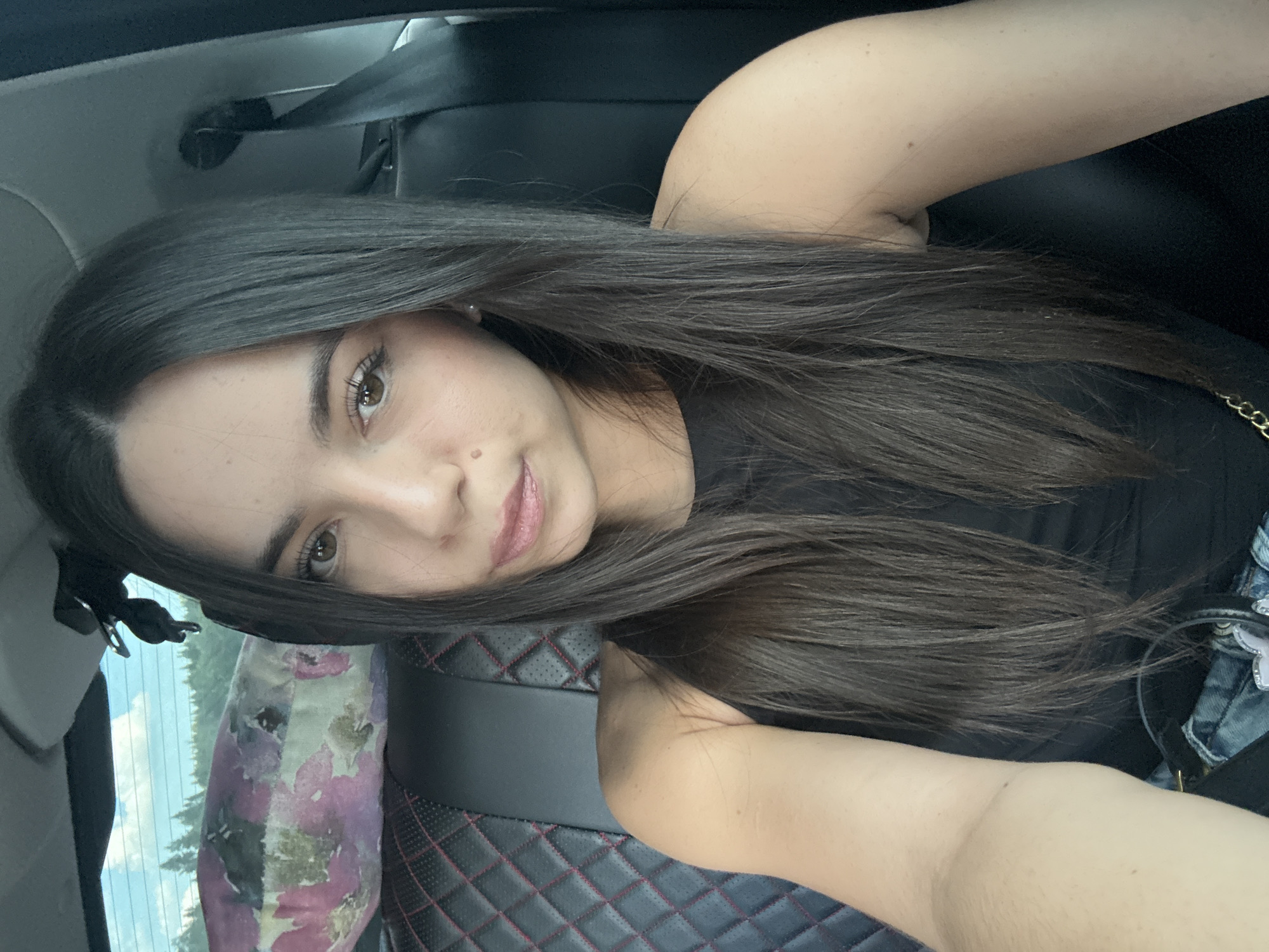 In a front-facing, indoor photograph taken from a low angle, a young woman with long, dark brown hair is seated in the passenger seat of a vehicle. She is wearing a black tank top and appears to be taking a selfie, as her arm is extended out of frame. Her gaze is directed at the camera, and her lips are slightly parted. The background shows a pillow with a floral pattern on the backseat of the car, and beyond that, a blurred view of trees and the sky through the rear window. The seat upholstery is black with red stitching in a diamond pattern. The scene is illuminated by natural light.
