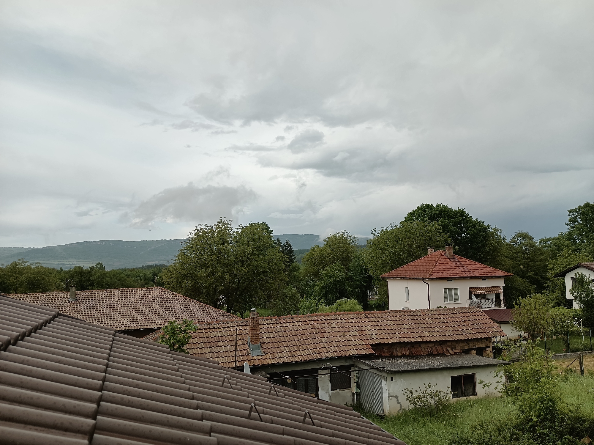 The image shows a view from above, likely a rooftop, in Mihaylovtsi, Bulgaria. In the foreground, a dark brown tiled roof dominates. Beyond this, several houses with red-tiled roofs are visible, interspersed with green trees. In the background, distant hills are visible under a cloudy, overcast sky. No people are visible in the image. The overall scene suggests a rural or suburban area. A small white structure is visible below one of the red-tiled roofs, partially obscured by vegetation.