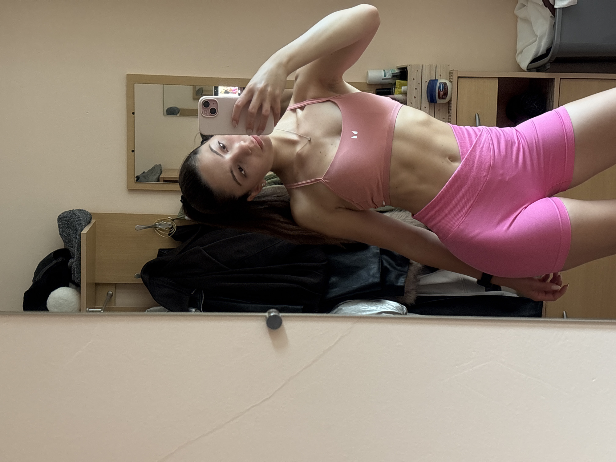 A young woman with dark hair pulled back in a ponytail is taking a mirror selfie in a room. She is wearing a pink sports bra and matching pink shorts. Her abs are visible, indicating she is fit. The background shows a closet with coats and shelves, a small mirror on a dresser, and a laundry basket. The room appears to be a bedroom or dressing area. The woman is holding a pink smartphone to take the picture. The lighting suggests it is daytime. No specific details are provided that would allow for identification of the location within the city of Varna, Bulgaria.
