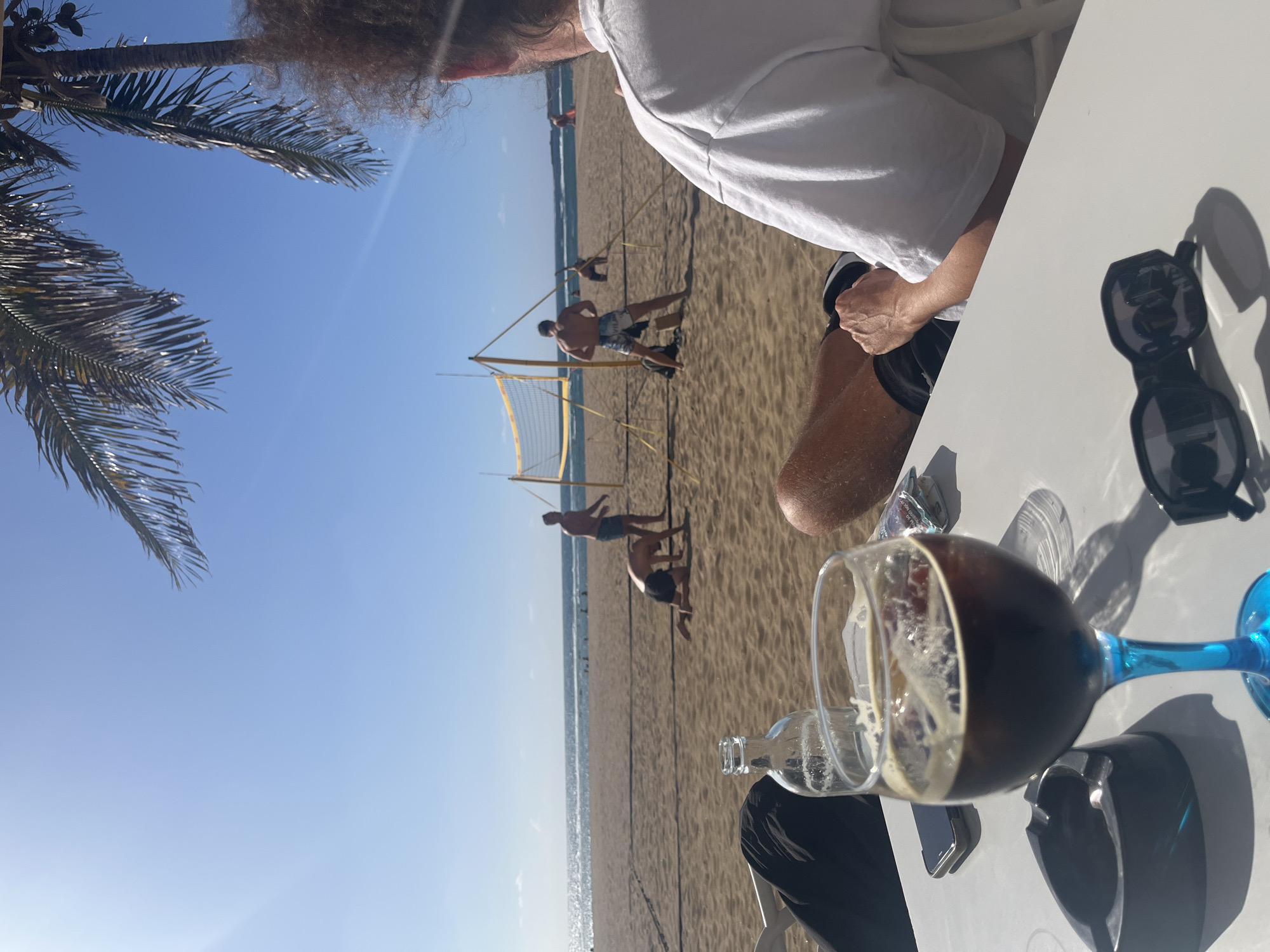 The image depicts a beach scene in Las Palmas de Gran Canaria, Spain, with a partial view of an individual seated at a table. In the background, a beach volleyball game is in progress, with several players near a yellow net. The beach stretches out to the sea under a clear blue sky. A palm tree frond is visible in the upper left corner. On the table, there is a dark-colored drink in a blue-stemmed glass, an empty bottle, sunglasses, a phone, and an ashtray. The individual at the table is wearing a white shirt.