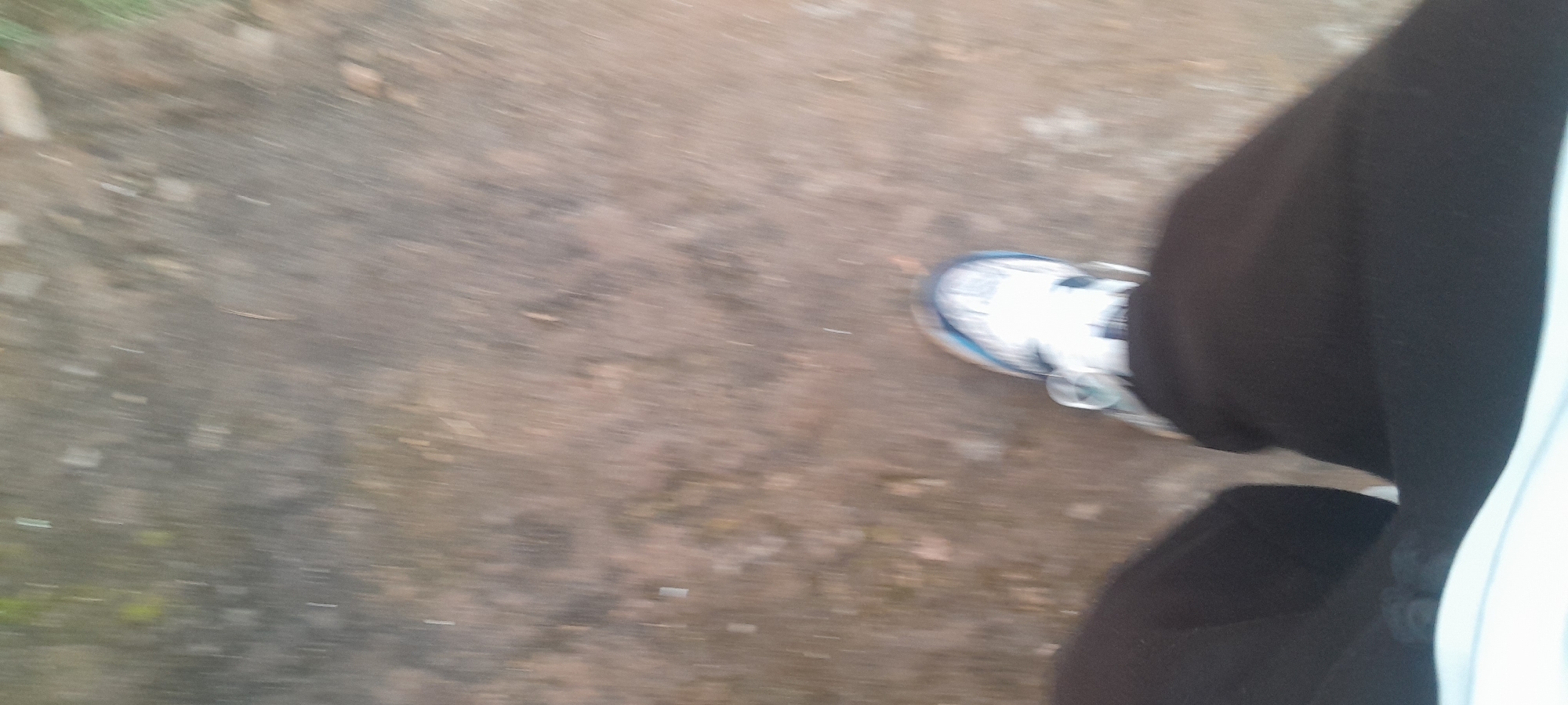 The image shows a person's leg and foot, wearing a white athletic shoe with blue accents and black trousers, walking on a rough, dirt or gravel path. The path appears to be outdoors, possibly in a park or undeveloped area. There are some small stones and debris on the ground. The background is out of focus due to motion blur, suggesting the photo was taken while walking. The lighting appears to be natural daylight. There are no visible landmarks to pinpoint the location to Abuja, Nigeria, or any specific city. The image captures a moment of movement on a simple outdoor surface.