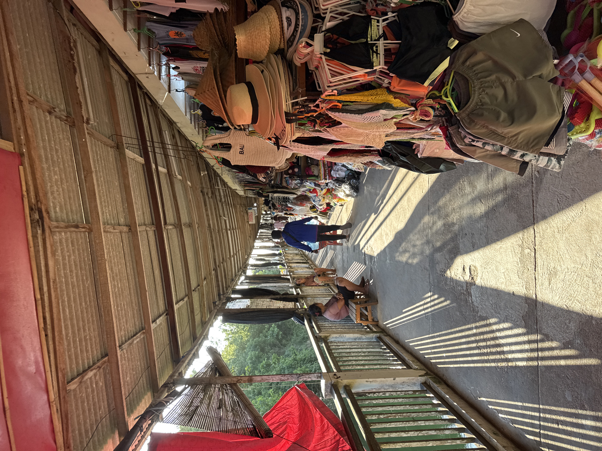 The image depicts a covered walkway, likely a market or bazaar, in Kuta Selatan, Indonesia. The walkway is lined with stalls displaying various merchandise, including hats, clothing (such as shorts with a Nike logo), and bags (one with "BALI" printed on it). The structure of the walkway appears to be made of corrugated metal and wood. People are visible walking through the walkway. One person is sitting on a small stool. The lighting is bright, with strong shadows cast across the ground, suggesting the time of day. The environment is commercial and populated.