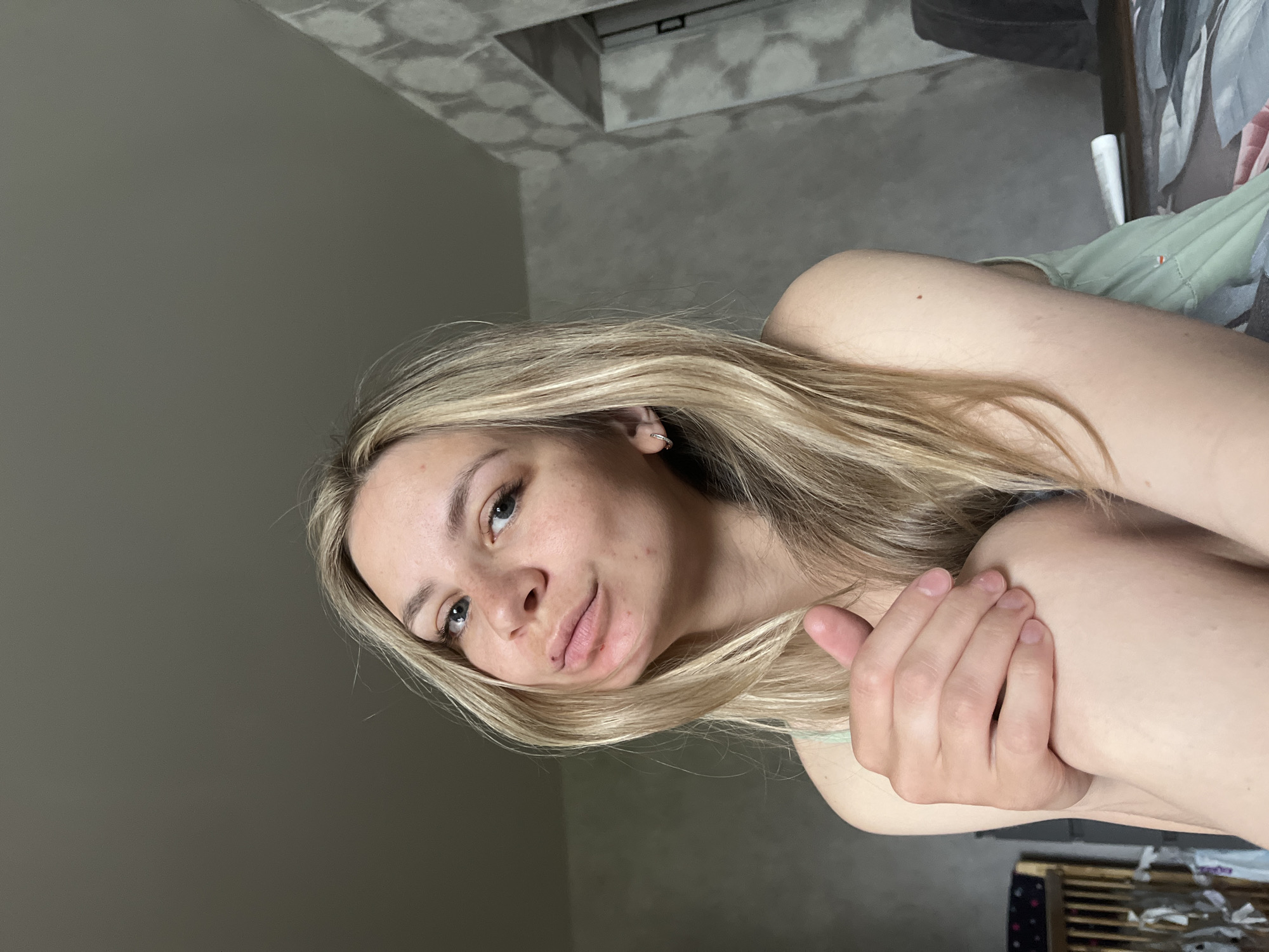 A woman with long blonde hair is photographed at a close range, holding her arm. The photograph was likely taken indoors, given the wall color and ceiling decor visible in the background. The location is in the city of Chaikovsky, Russia.