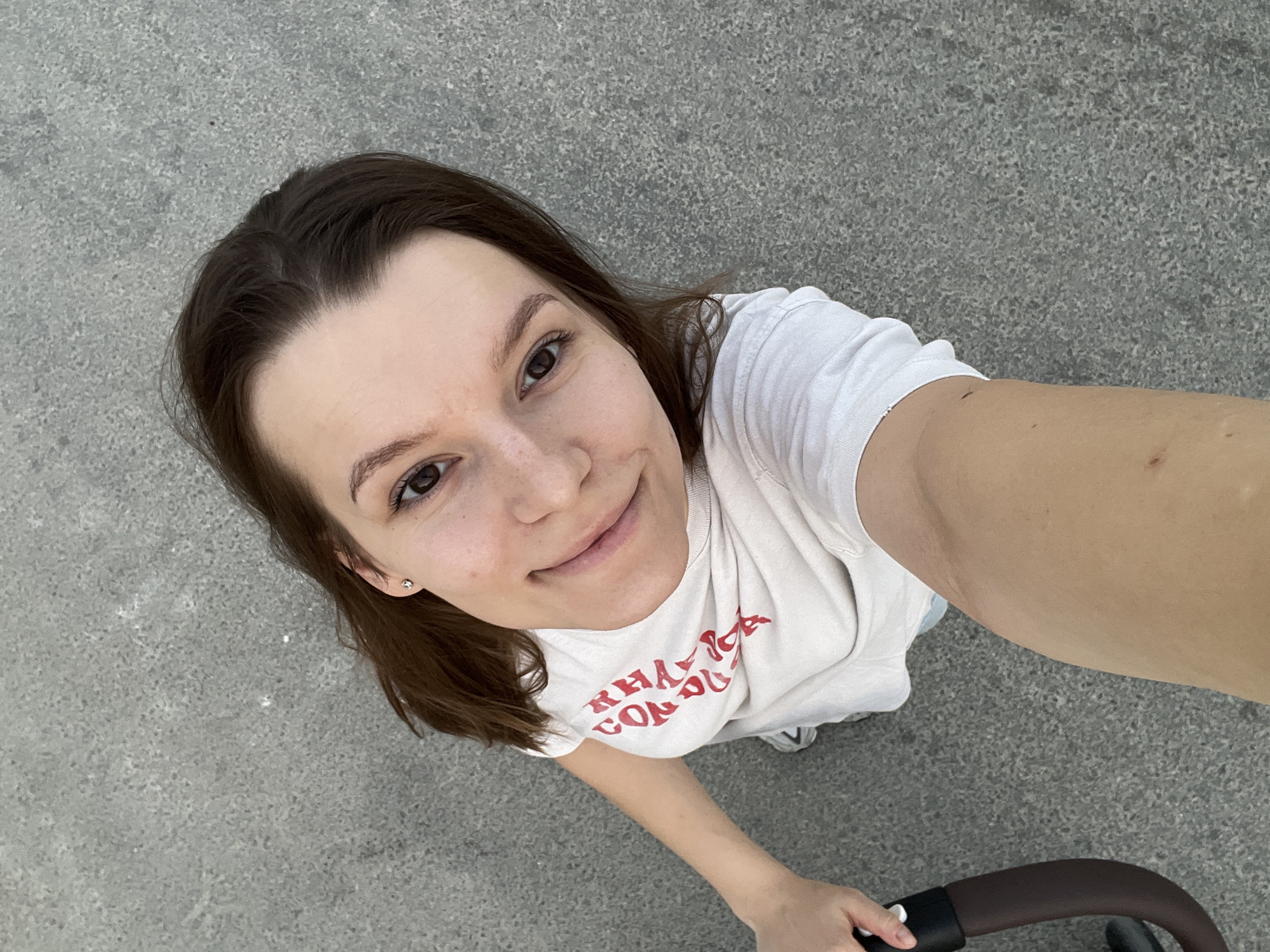 The image shows a young woman taking a selfie outdoors. She is wearing a white t-shirt with red lettering. She is holding onto a dark brown handle, possibly from a stroller or bicycle. The background is a gray, textured surface, likely pavement. The image is cropped closely to the woman's face and upper body. The lighting suggests it is daytime. No specific Limassol landmarks or identifying features are visible.