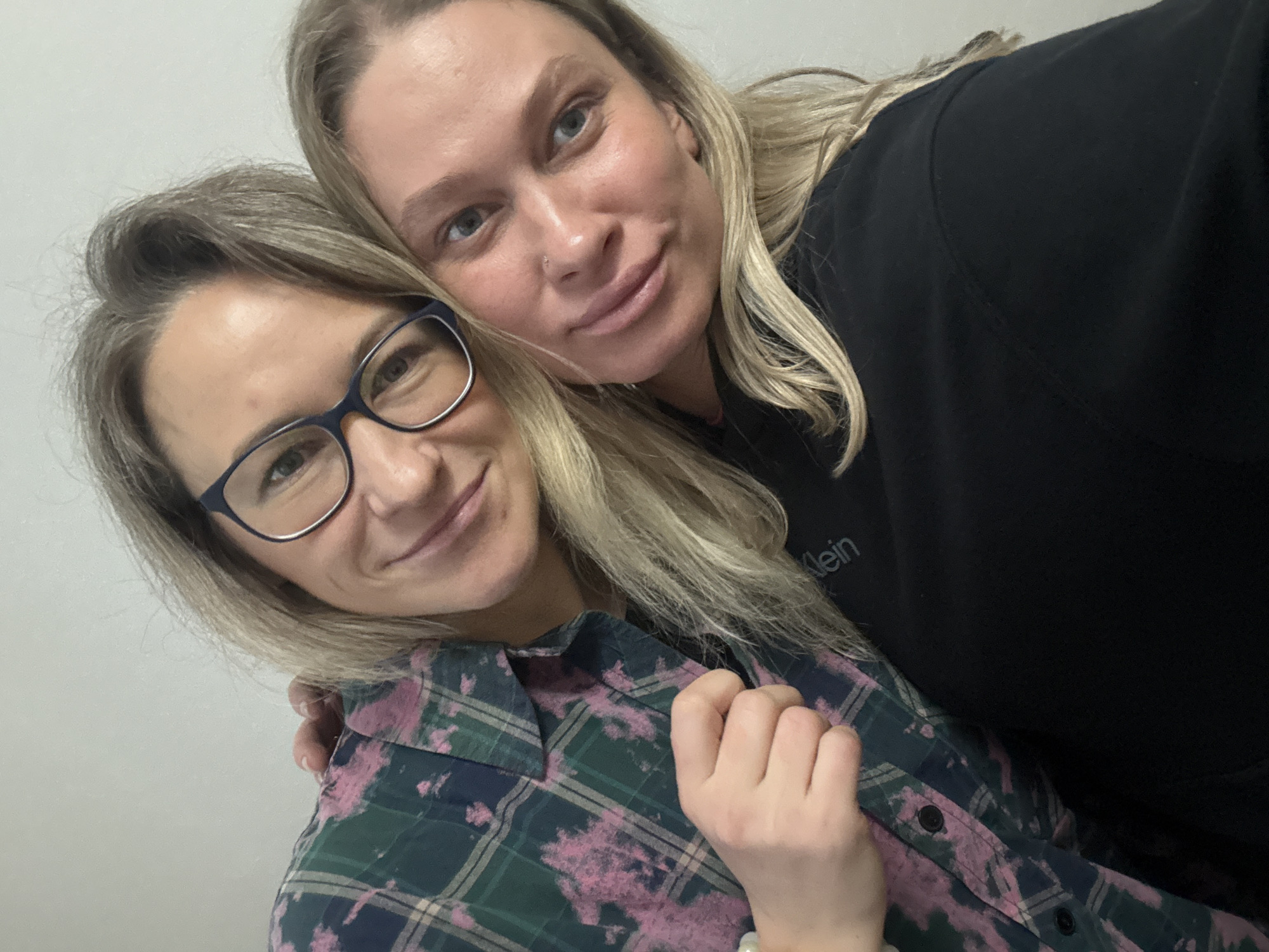 Two light-skinned women pose closely together indoors against a plain white wall. The woman on the left wears eyeglasses and a green, blue, and pink plaid shirt. The woman on the right wears a black top with the word "Klein" visible on it. The location of Кондратово, Russia is not visible in the image.